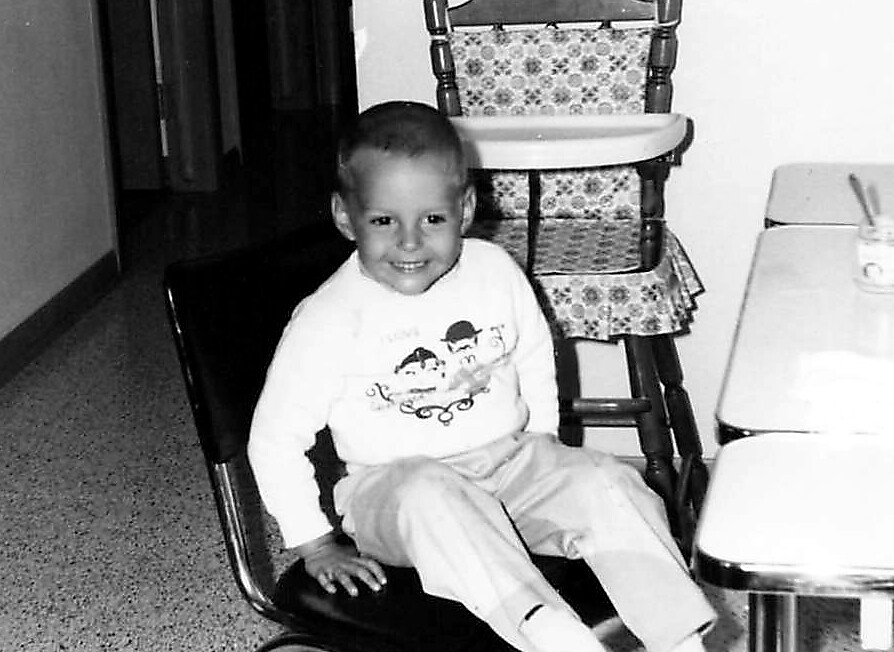 Smiling young child sits on a chair in a kitchen, with a high chair and small table nearby.