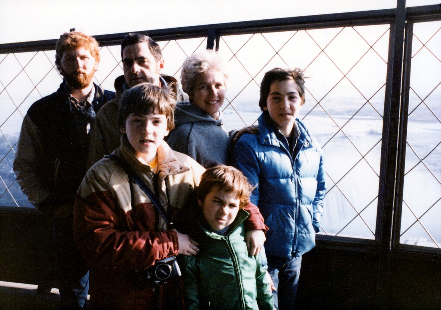Group of six people posing together on a high viewpoint, snow-covered landscape visible through a lattice-fenced window behind them, wearing warm jackets.