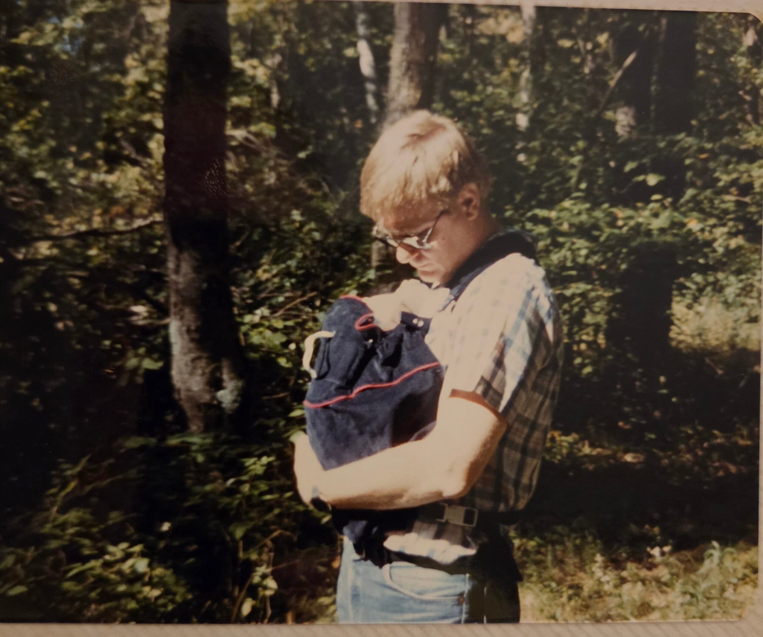 Person with blond hair wearing a plaid shirt and jeans stands in a wooded outdoor area.