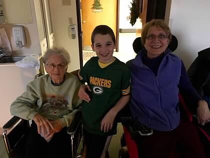 Three smiling people posing for a photo: an elderly woman in a wheelchair on the left, a boy in a Green Bay Packers shirt in the middle, and a woman in a purple jacket on the right, in a hallway.