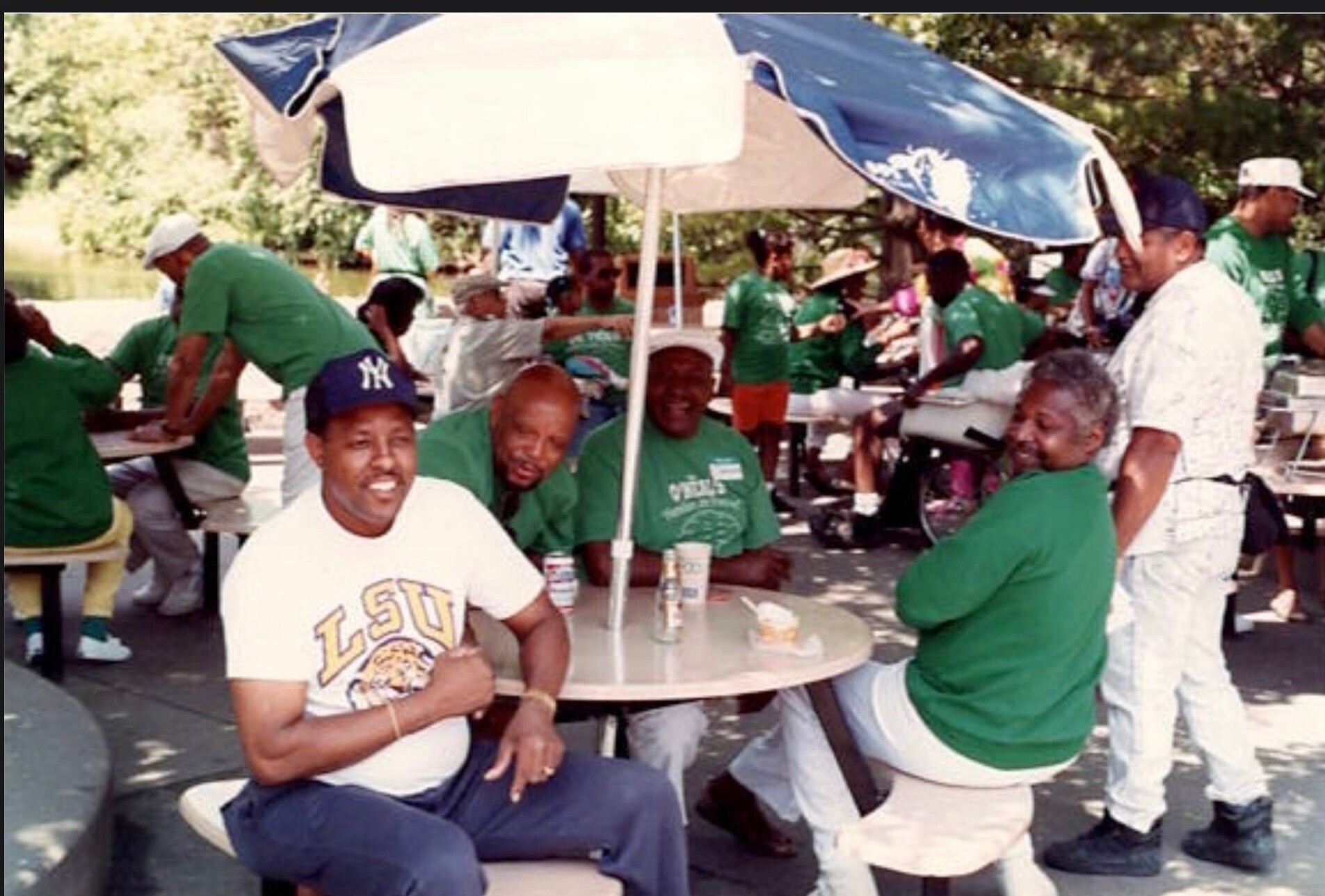 Group of people at an outdoor community picnic under a blue canopy, many wearing green shirts and sitting at tables.