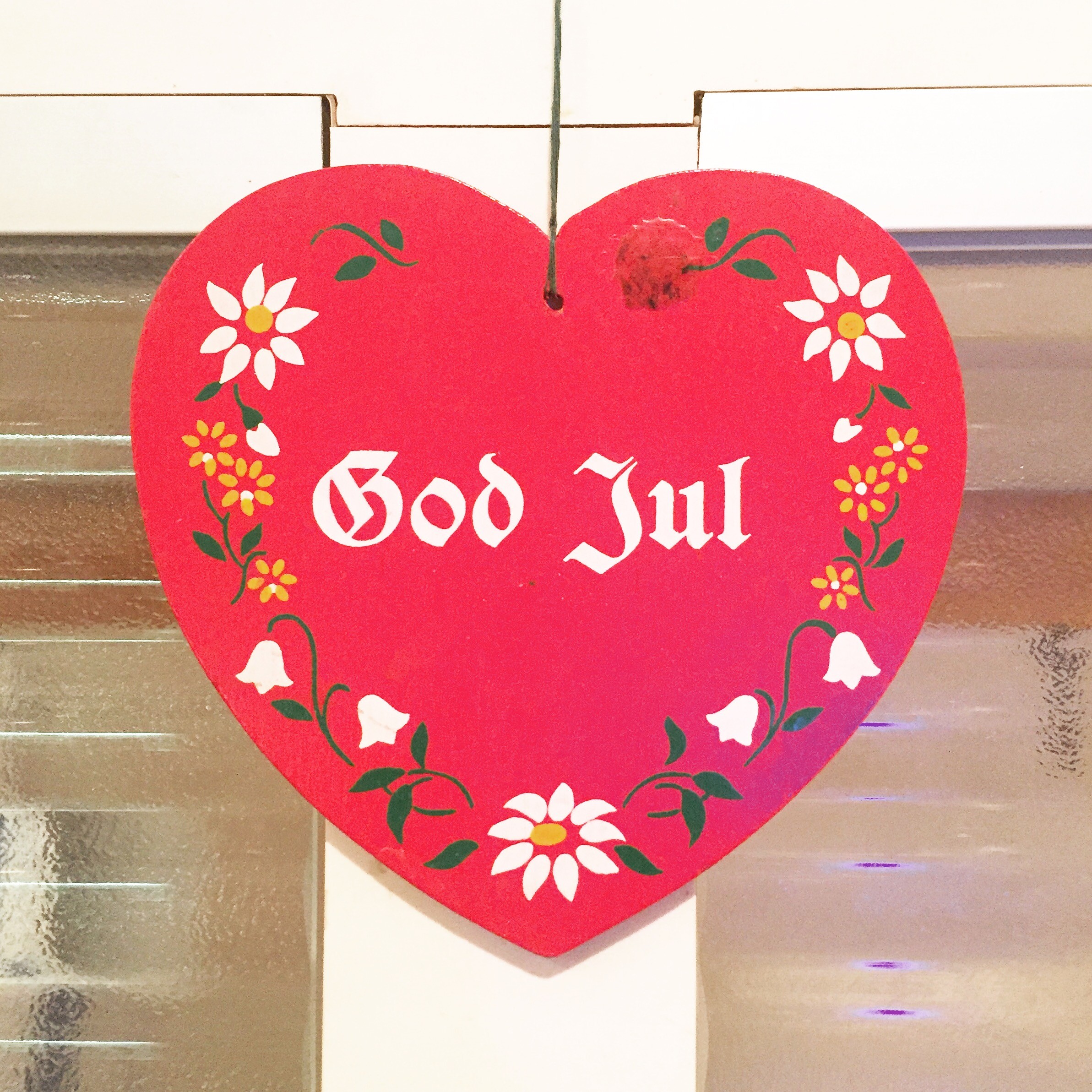Red circular Christmas sign with white Gothic text 'God Jul' and floral border, hanging from a string.
