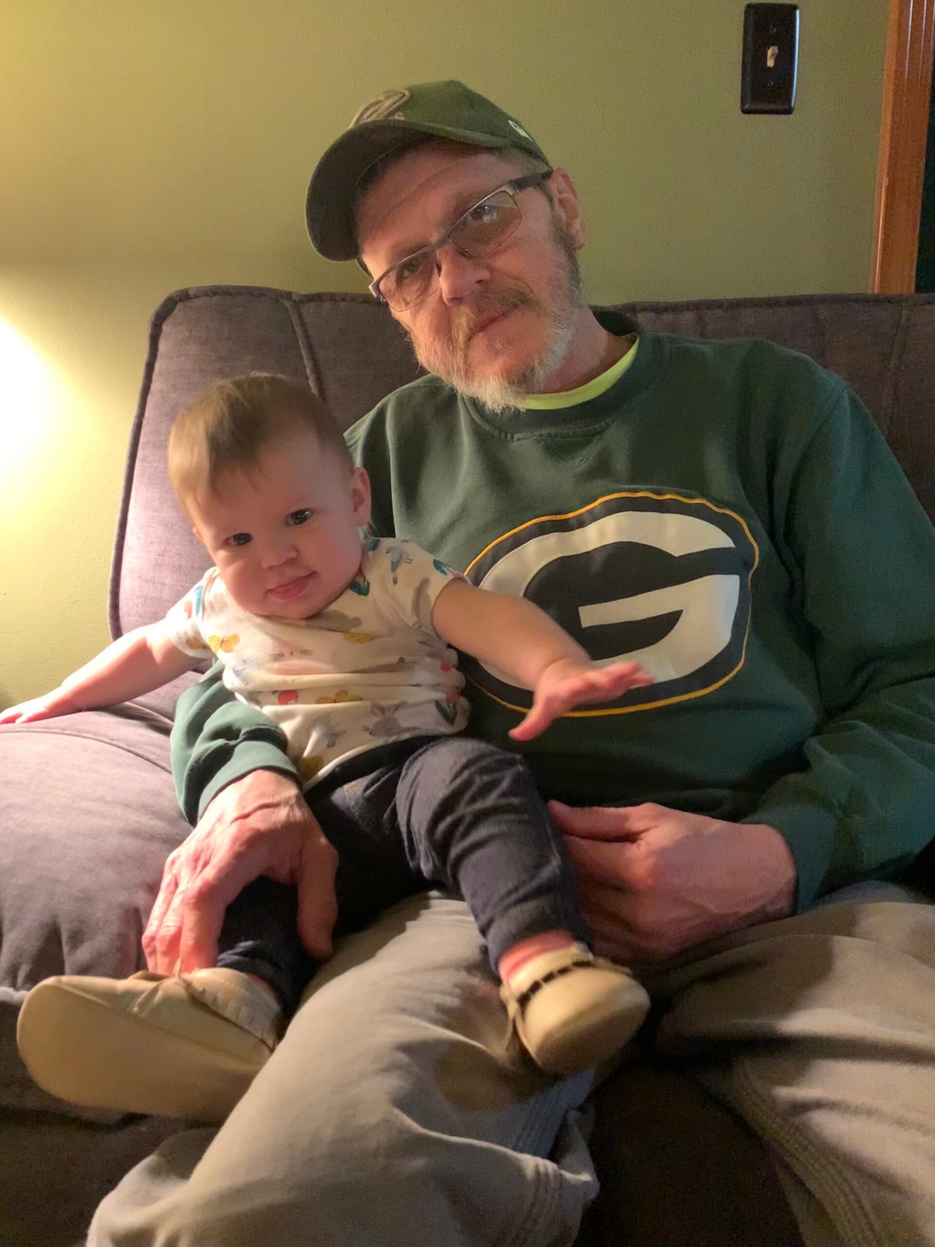 Older man with a green cap and glasses sits on a couch, holding a baby in a patterned onesie.