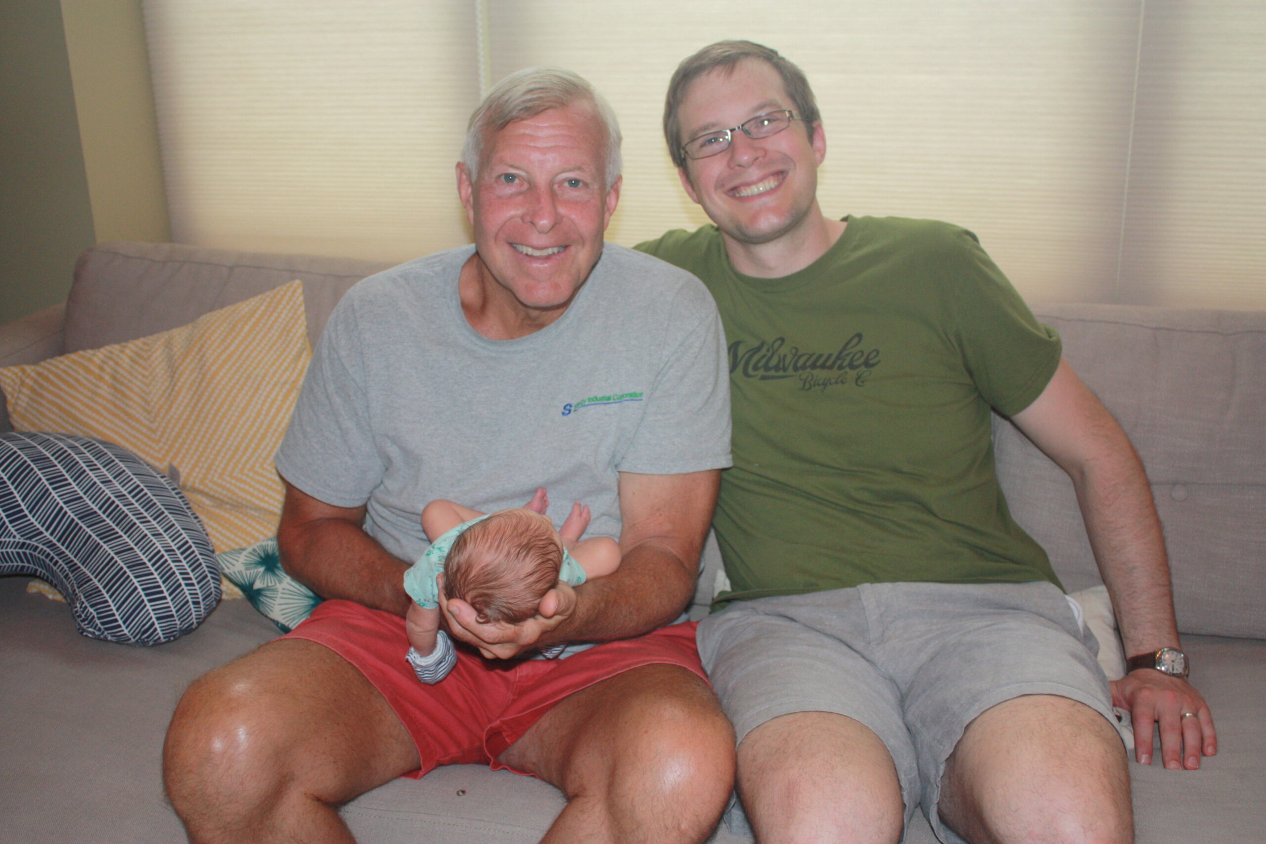 Two men sit on a sofa with a newborn baby cradled in the older man's arms, both smiling at the camera.