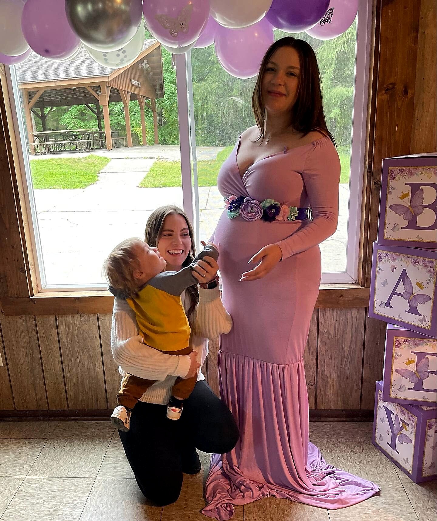 Pregnant woman in a pink dress with a floral belt stands next to a smiling caregiver holding a toddler indoors with pink balloons overhead.