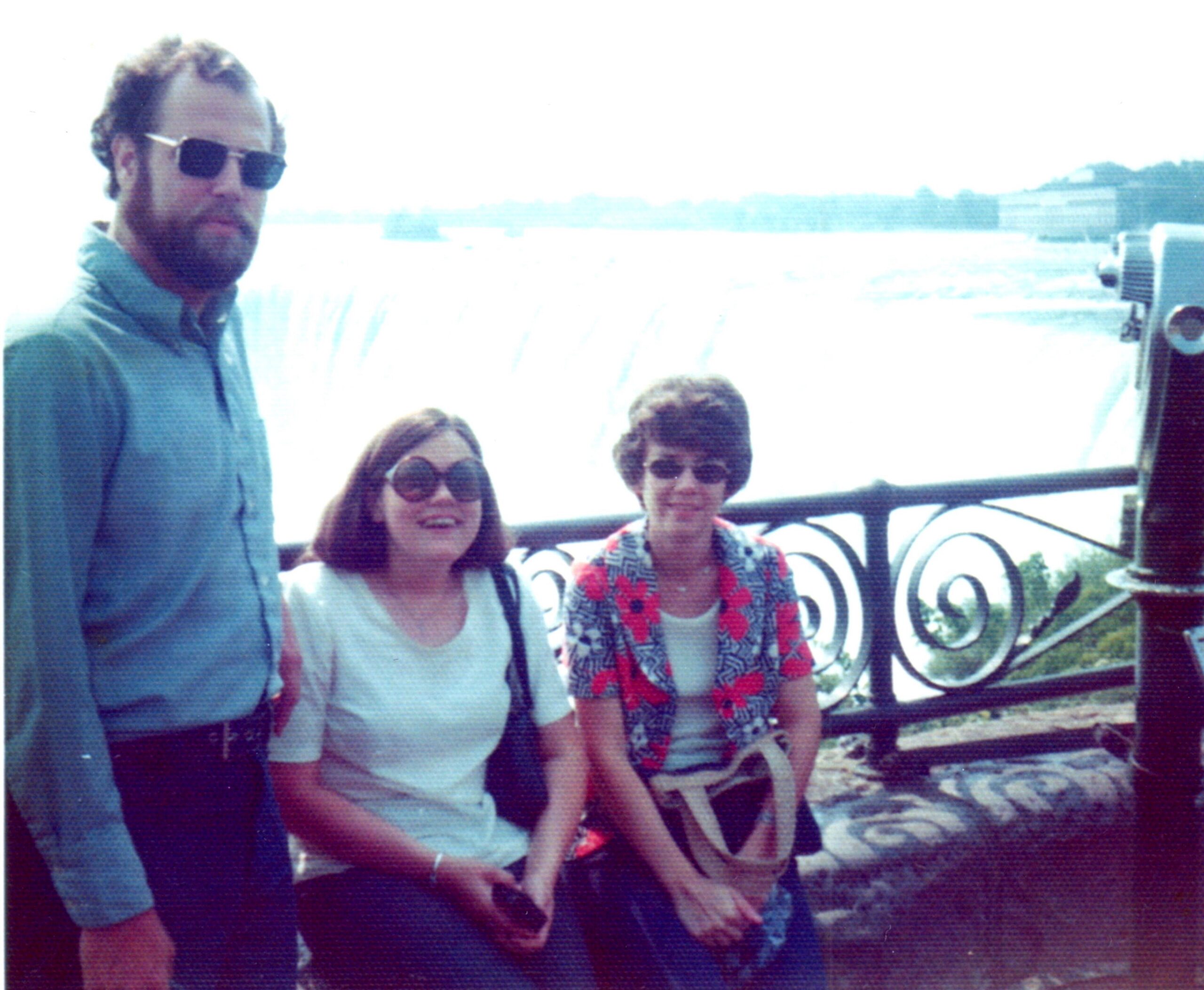 Four adults posing together outdoors, wearing sunglasses, with a decorative black-and-white railing in the background on a sunny day.