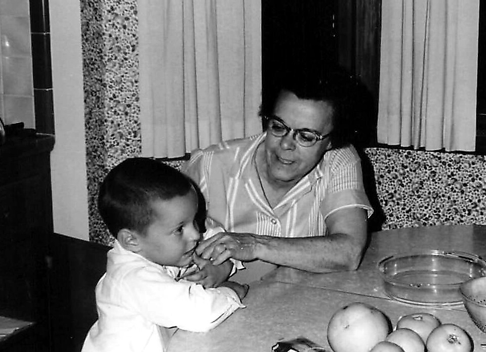 Woman with glasses sits at a kitchen table with a young child, hands touching as they interact; apples and bowls on the table are visible.
