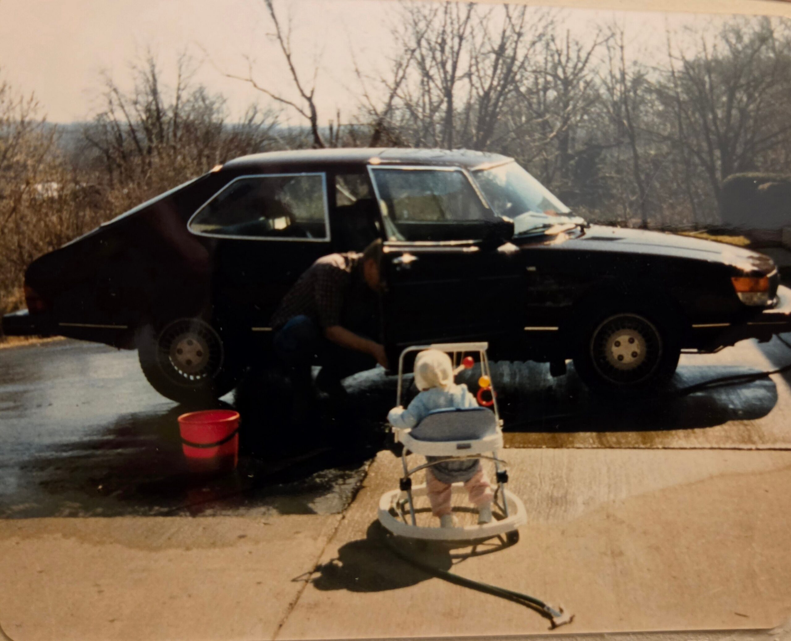 Black car parked on a driveway with a small child in a white stroller in the foreground; red bucket nearby as trees are bare in the background.