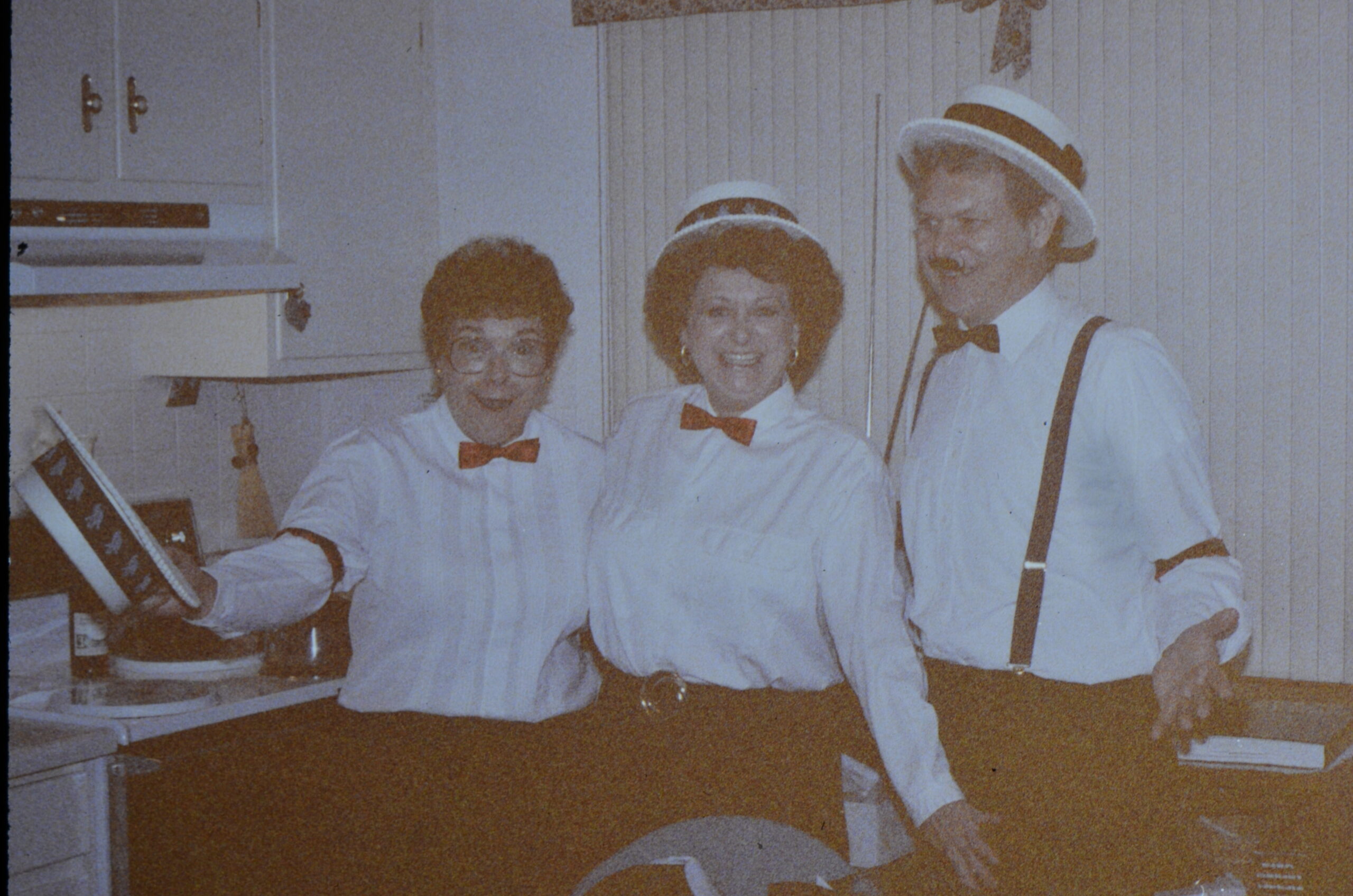 Three people in vintage outfits with red bow ties posing in a kitchen, smiling at the camera; left person holds a bottle, center wears a hat, right wears suspenders and a straw hat.
