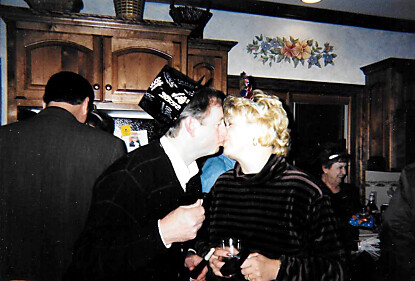 Two people kiss at a social gathering, holding drinks, in a kitchen with wooden cabinets and floral decor behind them.