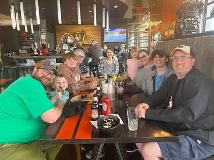 Group of friends and family gathered around a long table in a casual restaurant, smiling at the camera during a shared meal.
