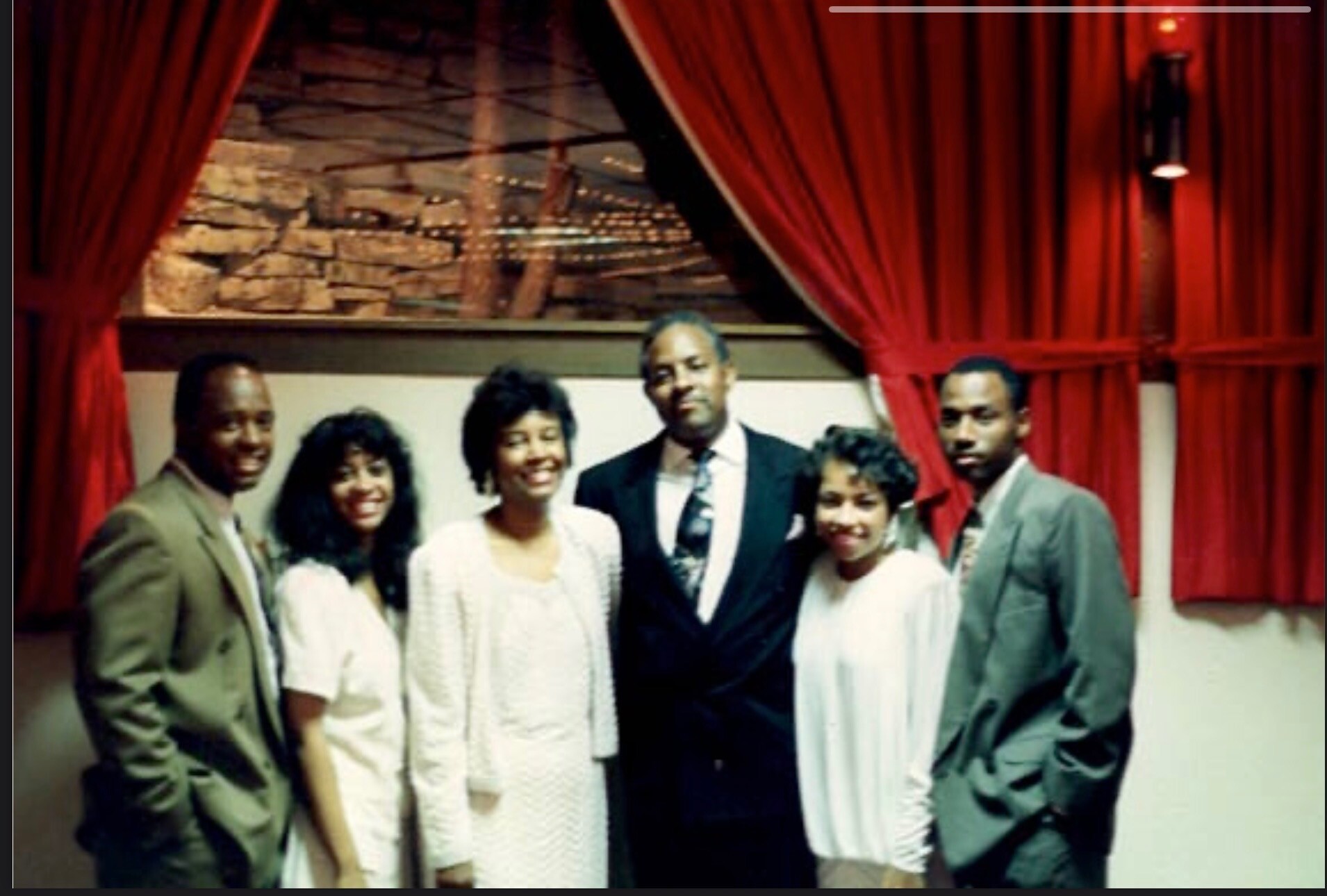 Group of six adults posing for a photo in a backstage area with red drapes and a stone wall, smiling at the camera.