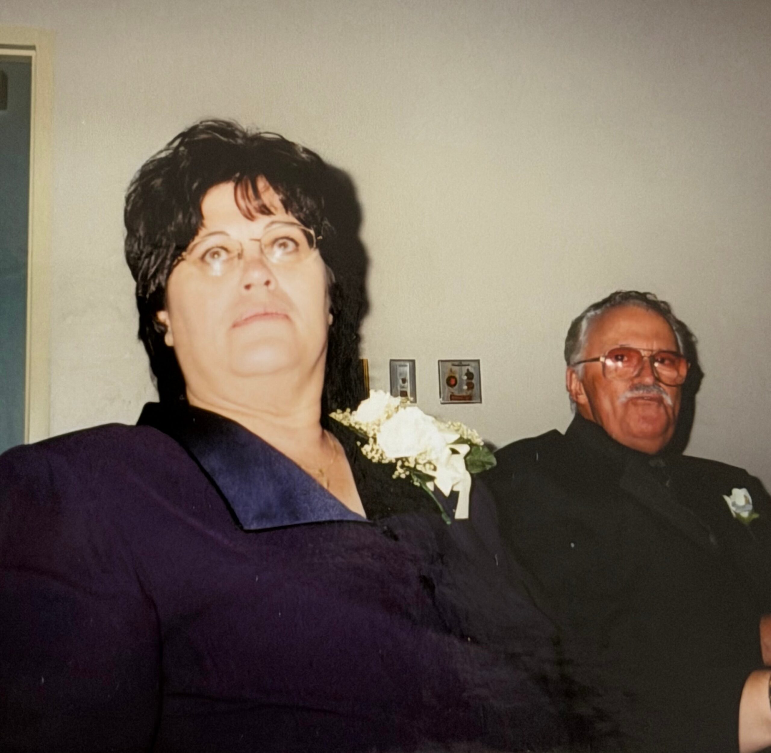 Woman in a dark purple outfit in the foreground, with a man wearing glasses behind her, indoors.