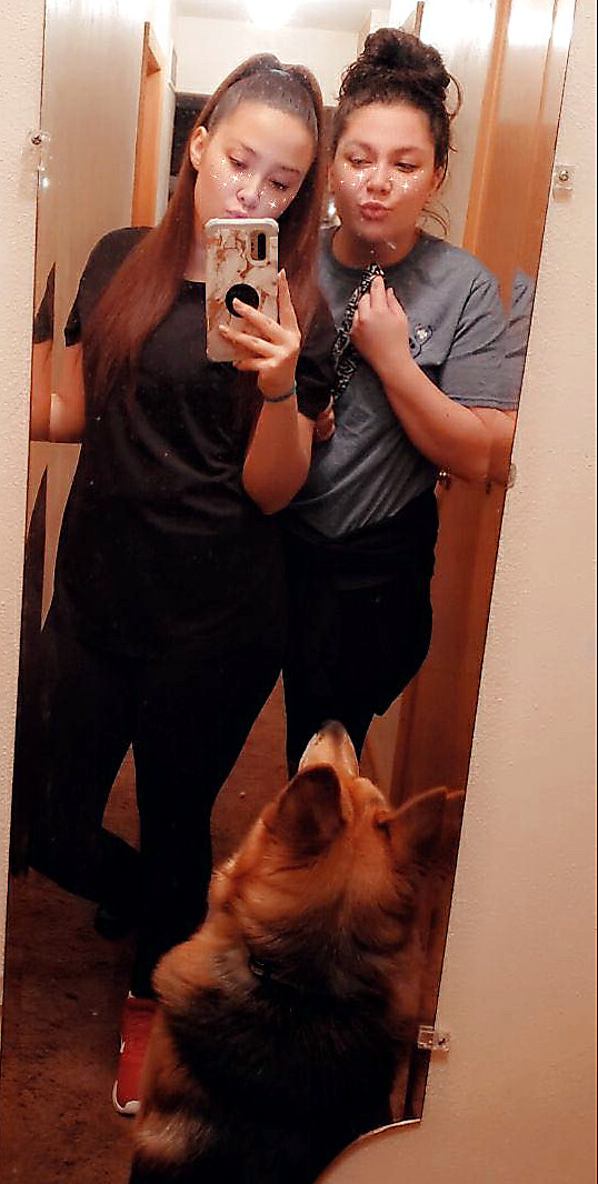 Two young women posing for a mirror selfie in a hallway, one holding a phone; a German Shepherd at the bottom looking up, both making pouty faces with a freckles filter.