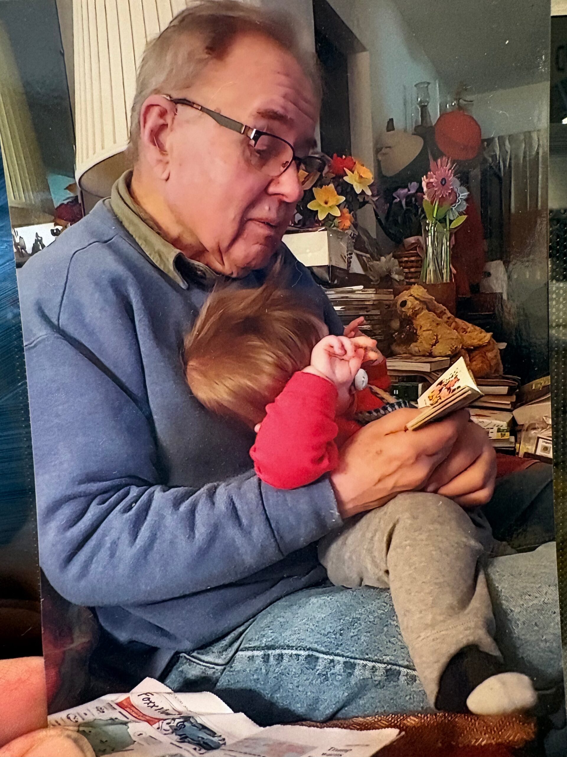 Elderly man in glasses holds a small child while reading a card in a cozy living room with flowers and books behind them.