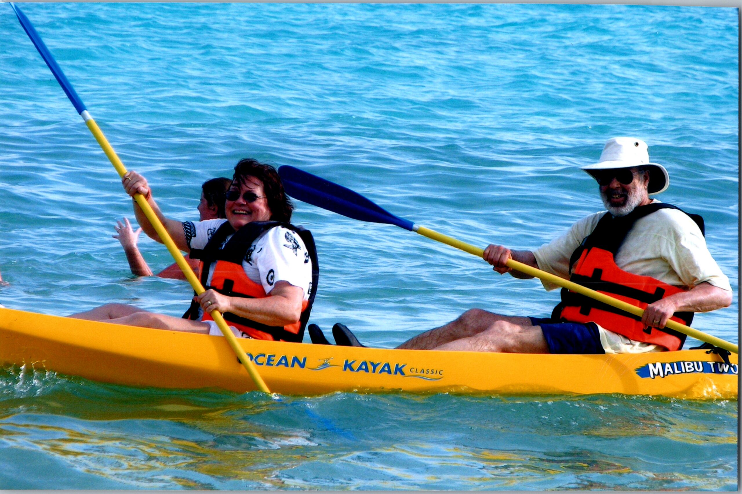 Two people paddling a yellow kayak on blue water, both wearing orange life jackets.