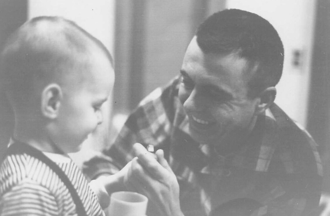 Man in a plaid shirt smiles at a baby who is holding a small object between fingers near a cup.