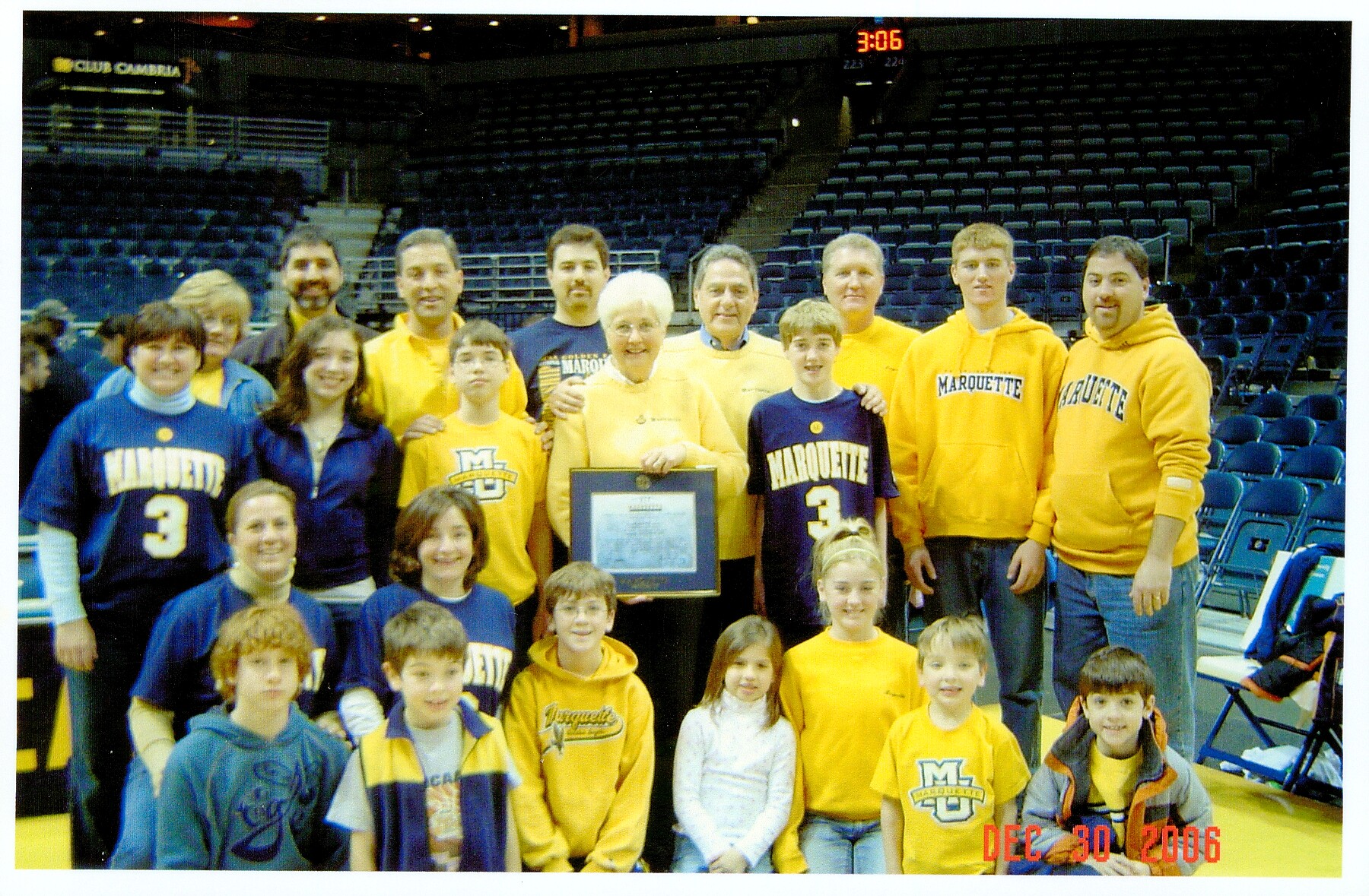 Group of kids and adults in a gym posing for a photo; several wear Marquette blue and gold, center woman holds a plaque/award.