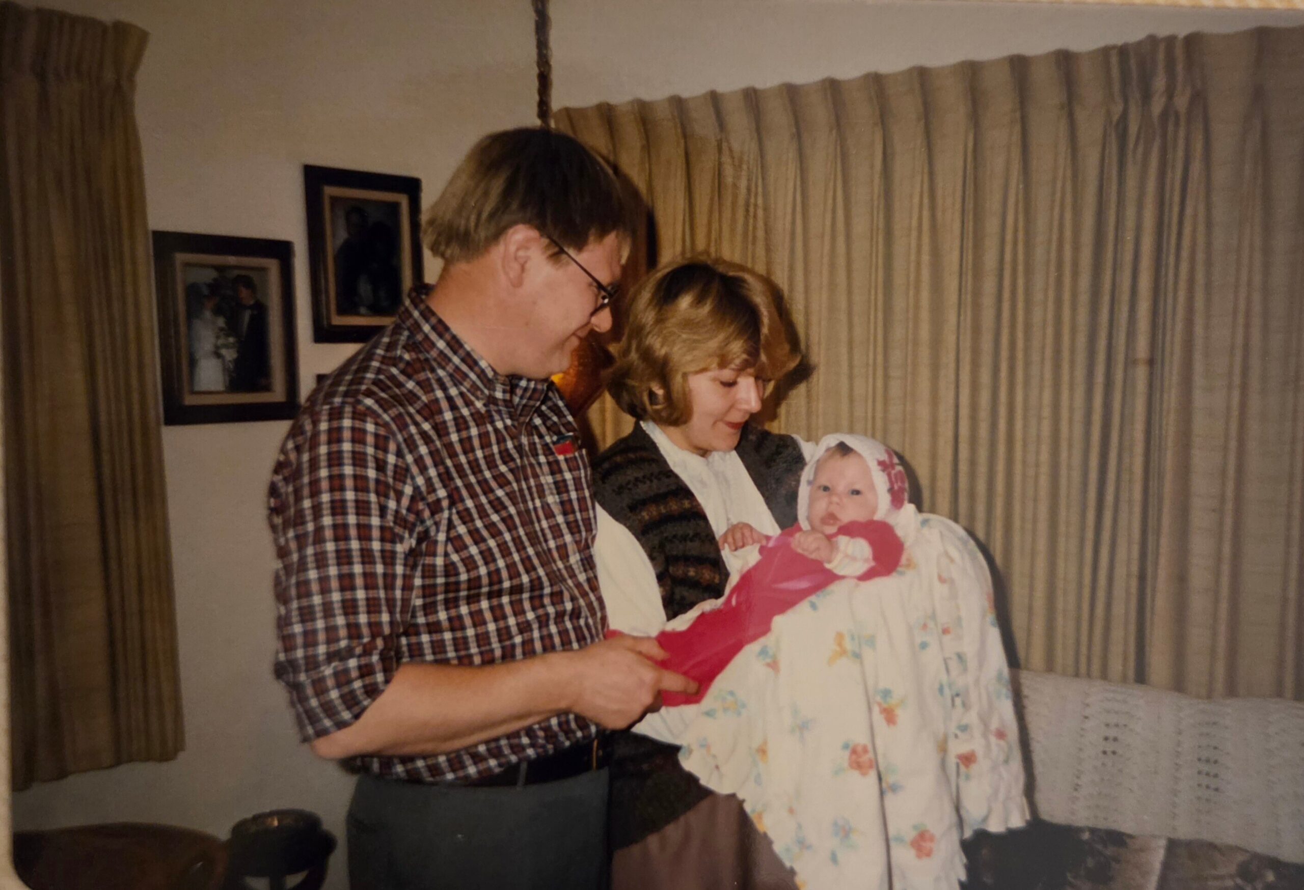 Older man wearing glasses holding a newborn swaddled in a floral blanket, with a woman beside them in a living room.