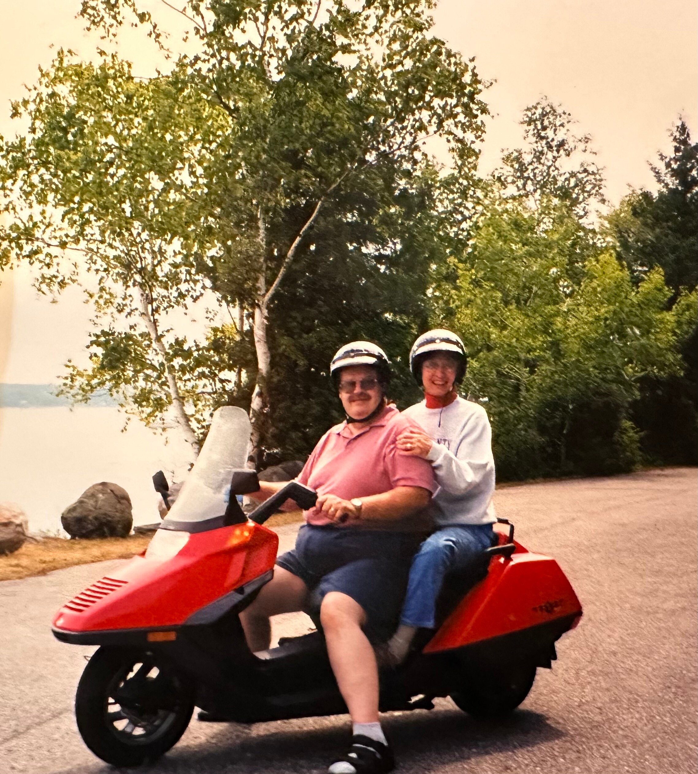 Two people wearing helmets ride a red motor scooter on a sunny day, posing with trees in the background.