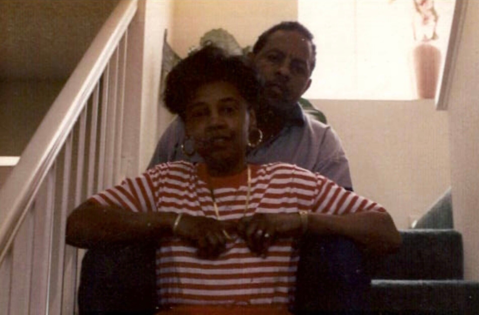 Two people pose on a staircase: a woman in a red-and-white striped shirt in front, a man behind her on the steps.