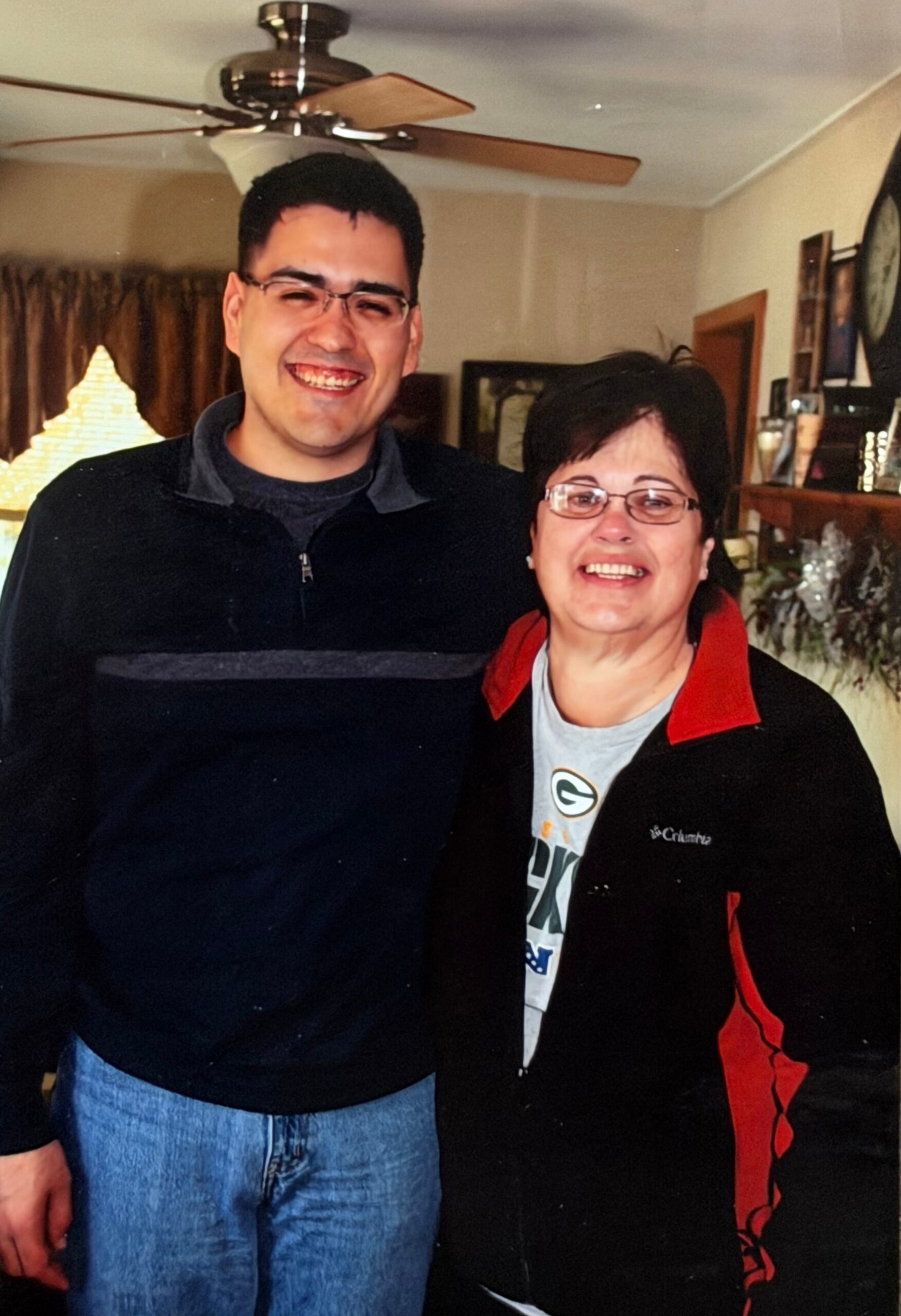 Smiling man and woman pose for a photo indoors in a living room, with a ceiling fan above them and shelves in the background.