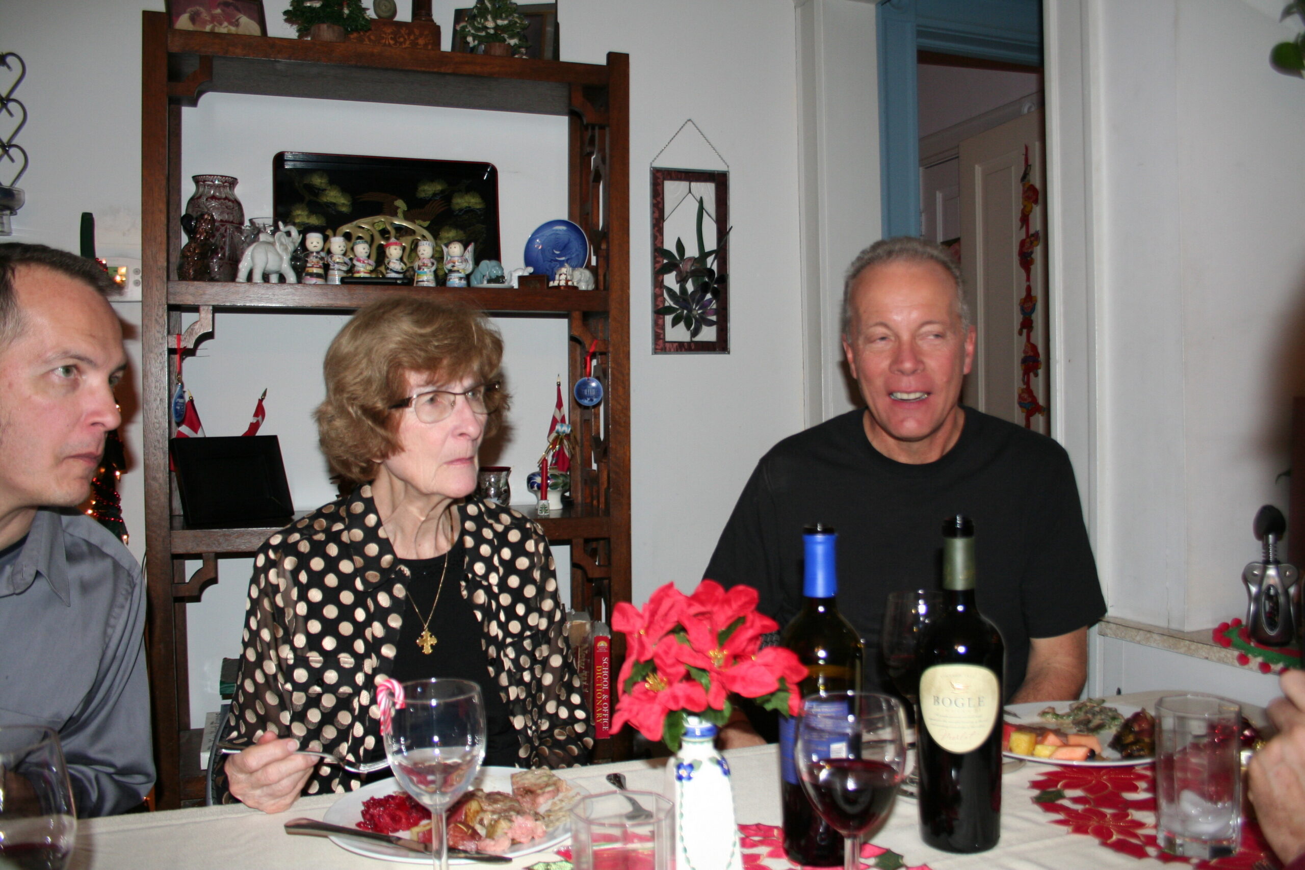 Three adults seated around a dining table with wine glasses and plates; red centerpiece flowers and a bottle of wine in foreground; a wooden shelf with figurines in the background.