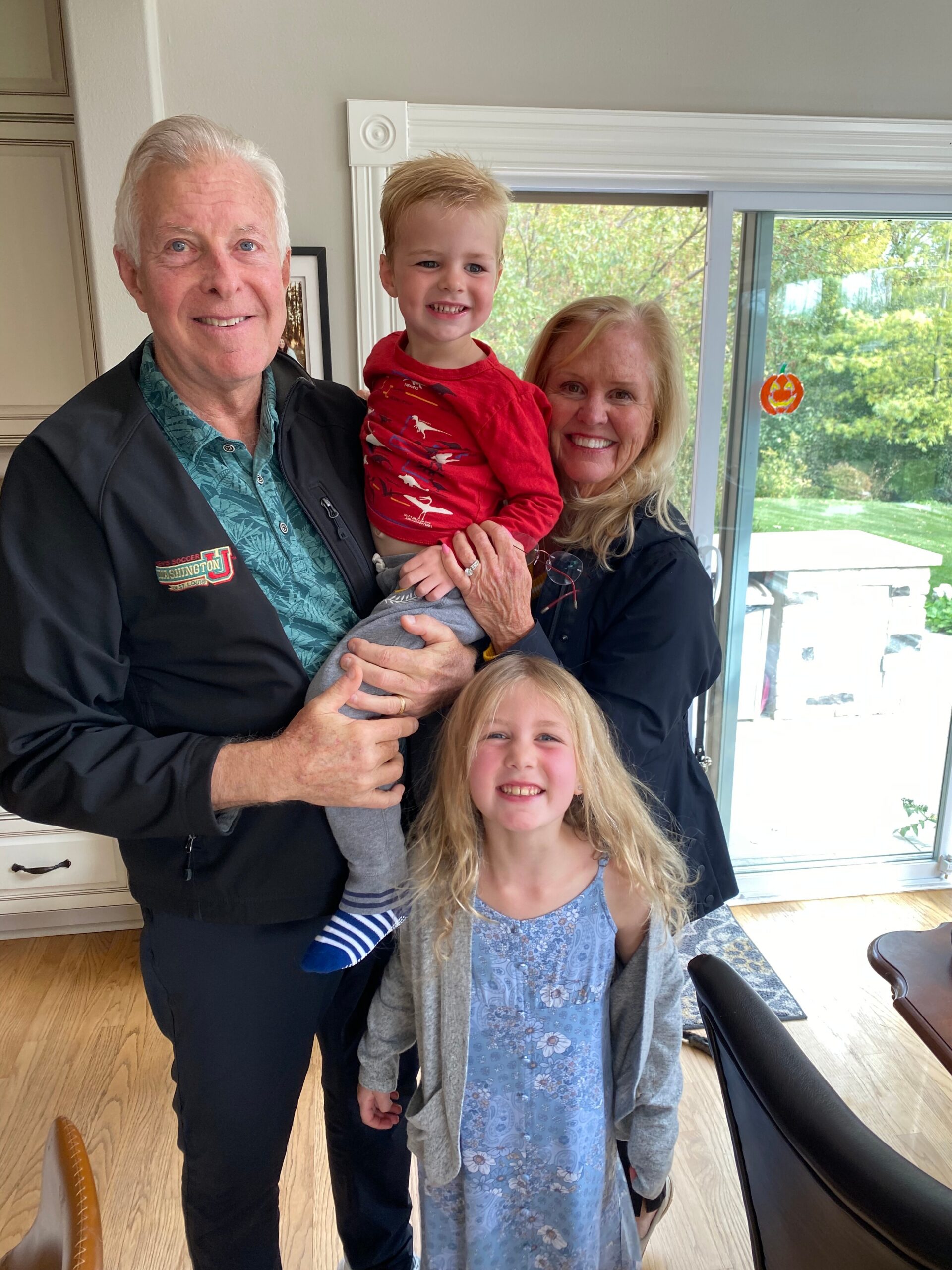 Grandfather holding a smiling toddler while his mother stands behind them by a sliding glass door in a bright kitchen.