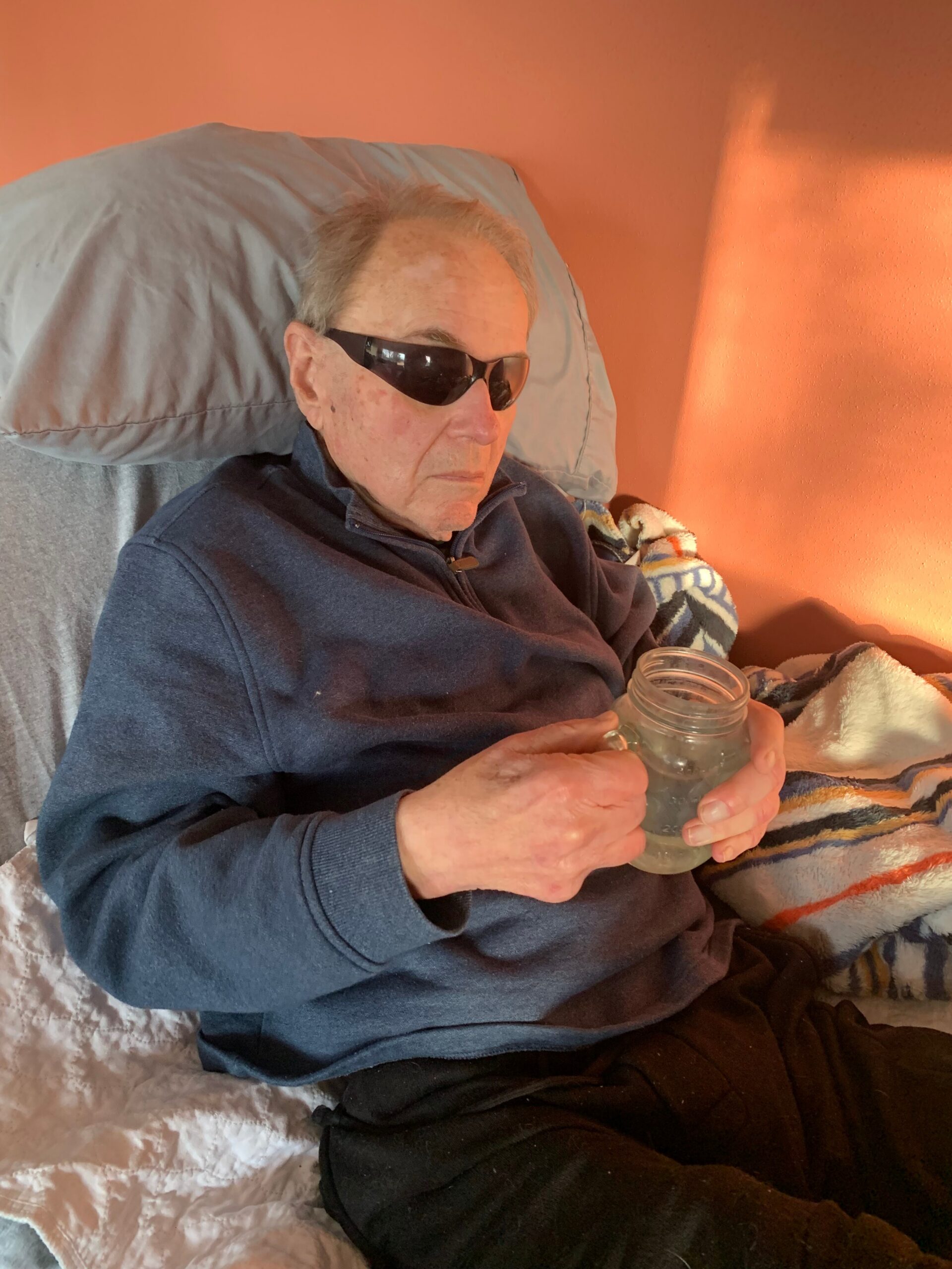 An elderly man wearing sunglasses sits in bed holding a glass jar with a light liquid inside, resting against a blue jacket and orange wall behind him.
