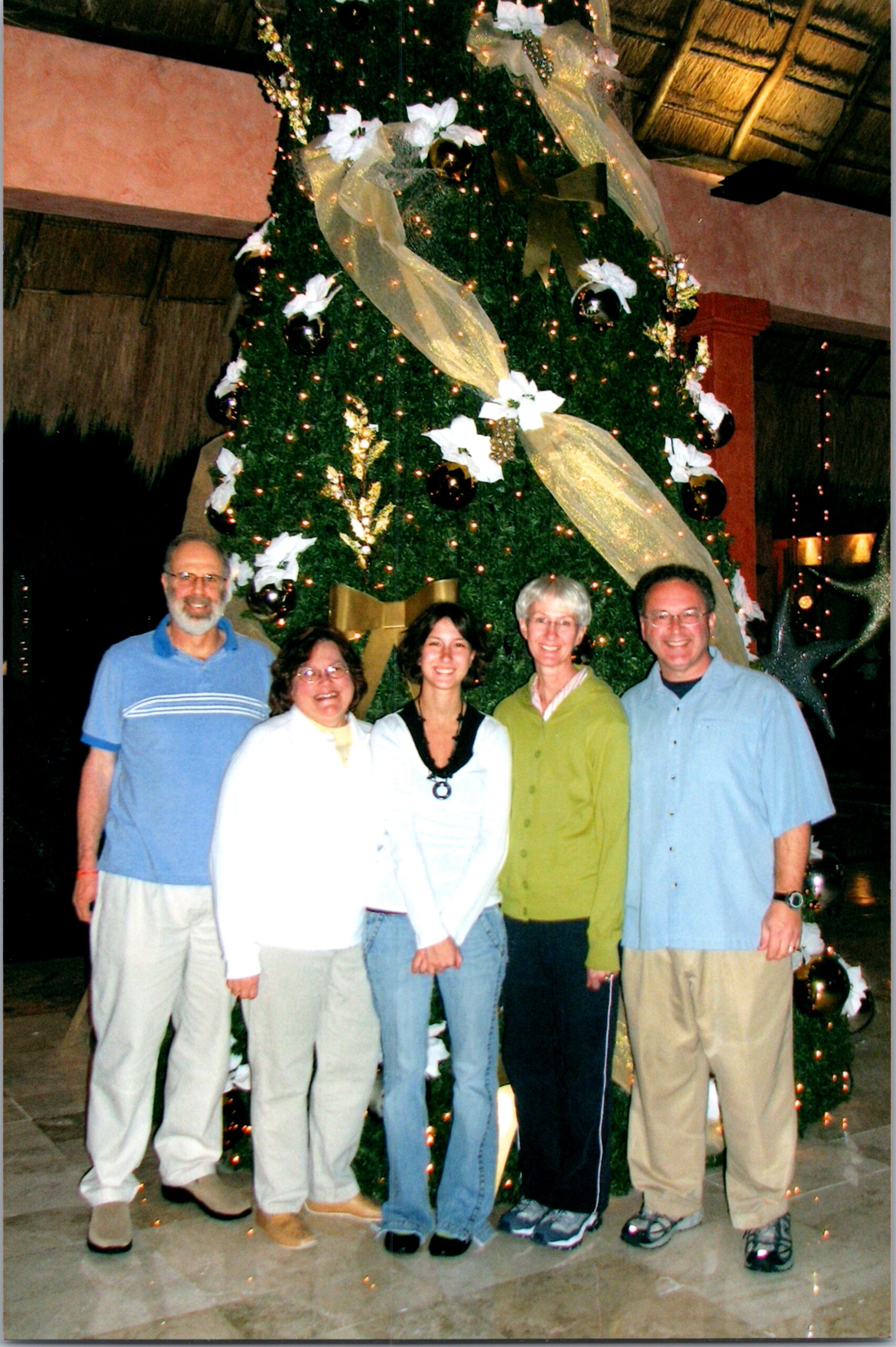 Six adults posing in front of a large, decorated Christmas tree indoors, smiling at the camera.