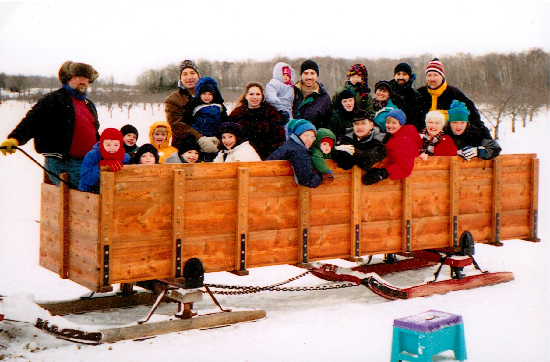 Group of adults and children posing on a large wooden wagon sled in a snowy field. A winter outing outdoors.