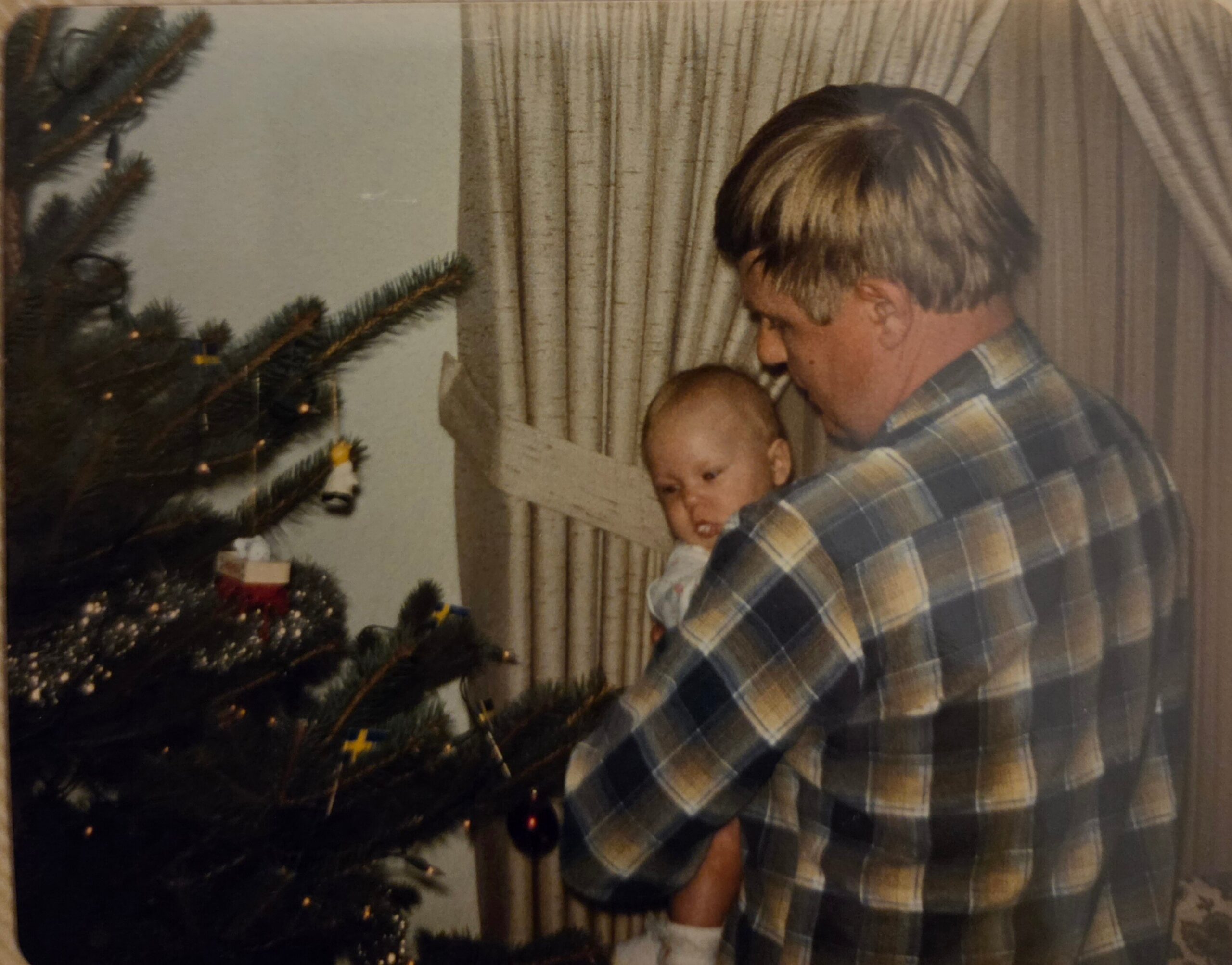 Man with blond hair wearing a plaid shirt holding a baby next to a lit Christmas tree at home.
