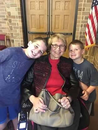 Smiling grandmother seated between two children, with arms around them, in a brick-walled room with a wooden door and an American flag nearby.