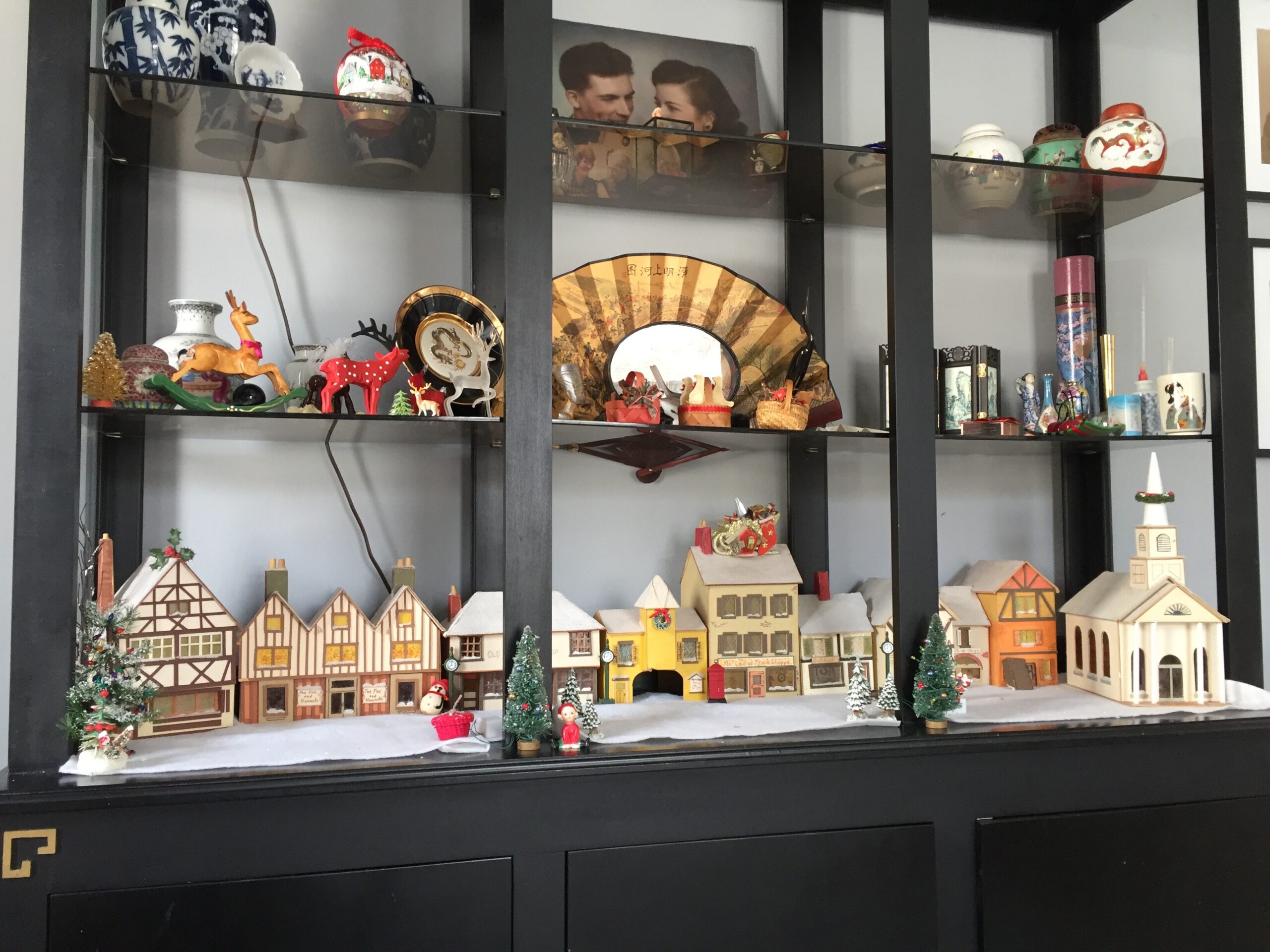 Shelf display of a miniature Christmas village with wooden houses, tiny trees, and festive figurines in a black frame cabinet.