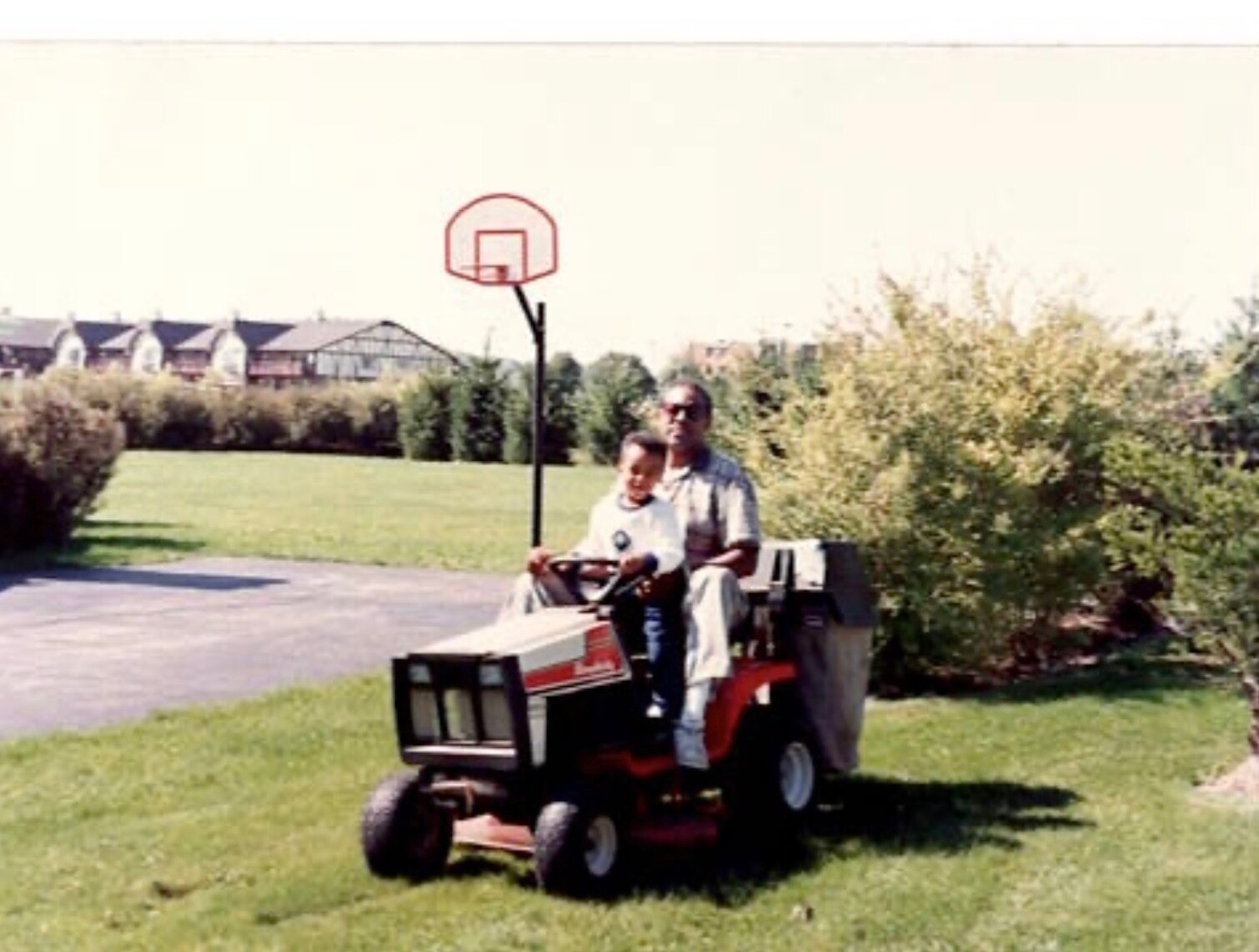 Man driving a red riding lawn mower with a child sitting on his lap, in a grassy yard with a basketball hoop and houses in the background.