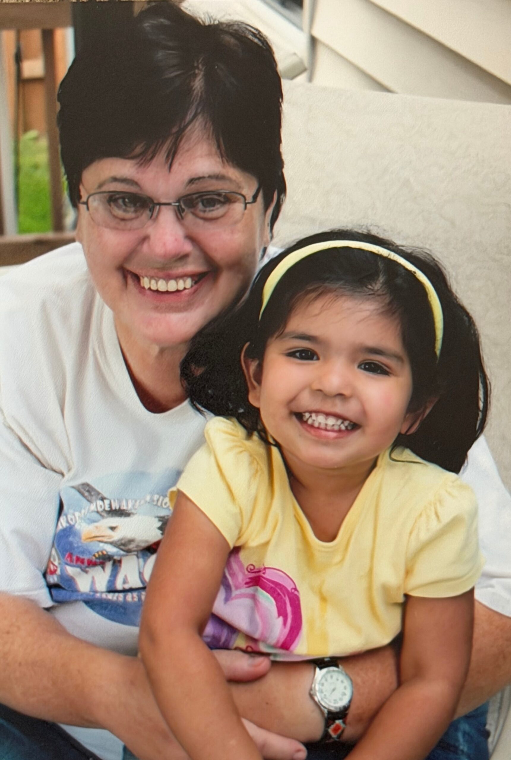 Smiling woman with short dark hair and glasses posing with a young girl wearing a yellow headband outdoors.