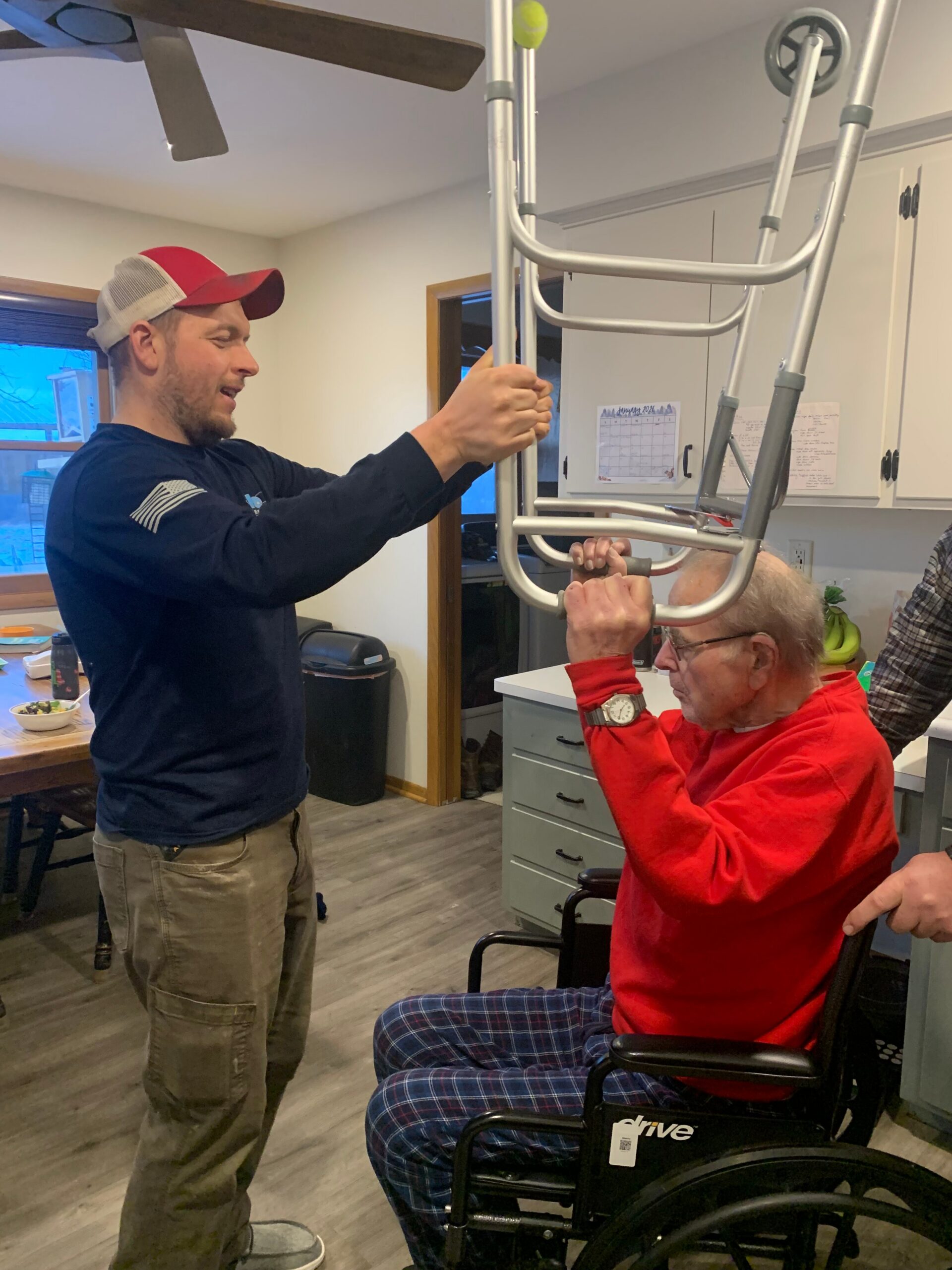 Two men help an elderly man in a wheelchair position a tall metal patient lift frame, preparing to assist with standing or transferring him.