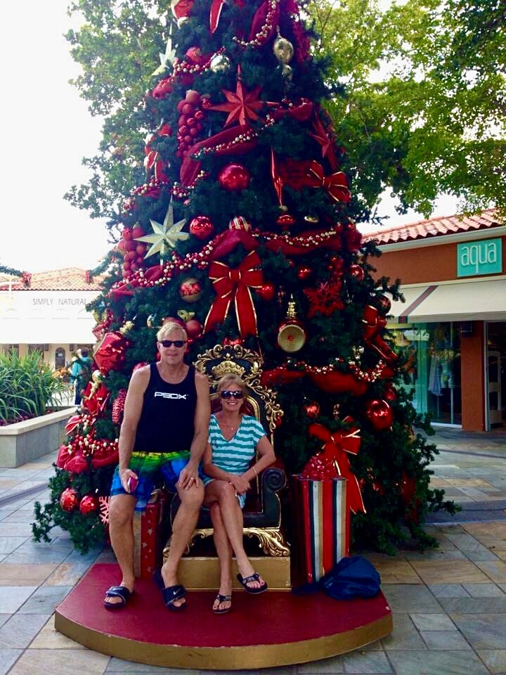 Two people sit on a gold ornate throne beside a gigantic Christmas tree decorated with red ribbons and ornaments in an outdoor space.