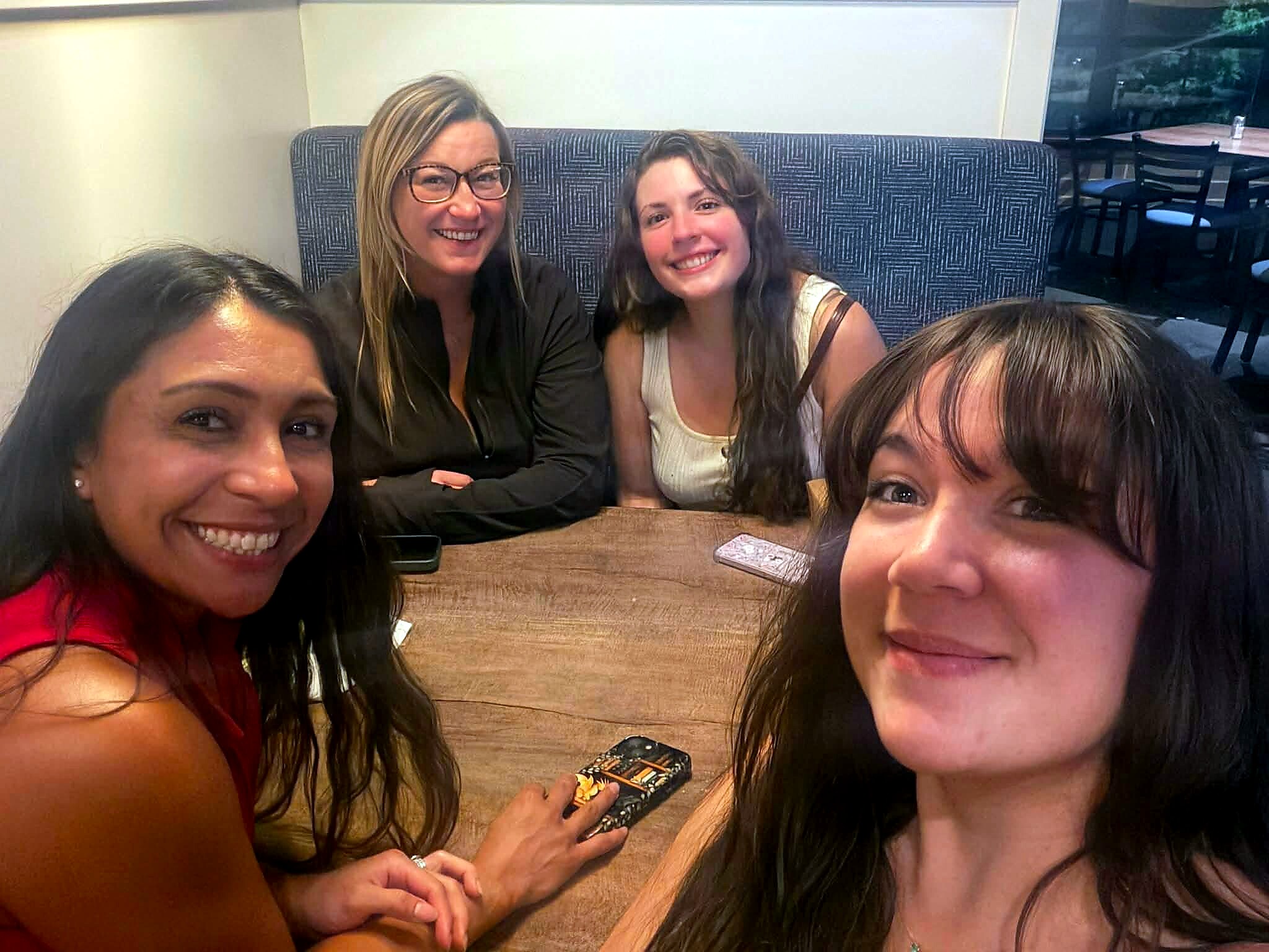 Four women smiling at the camera while posing for a selfie at a casual restaurant table.