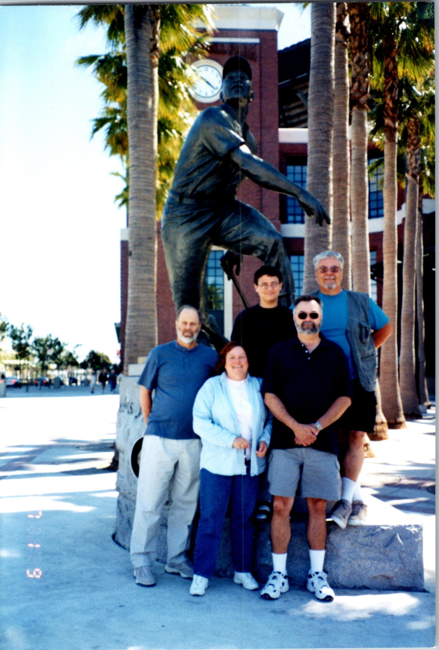 Group of six people posing for a photo in front of a large bronze statue of a kneeling man, outdoors among palm trees with a brick building and clock tower in the background.