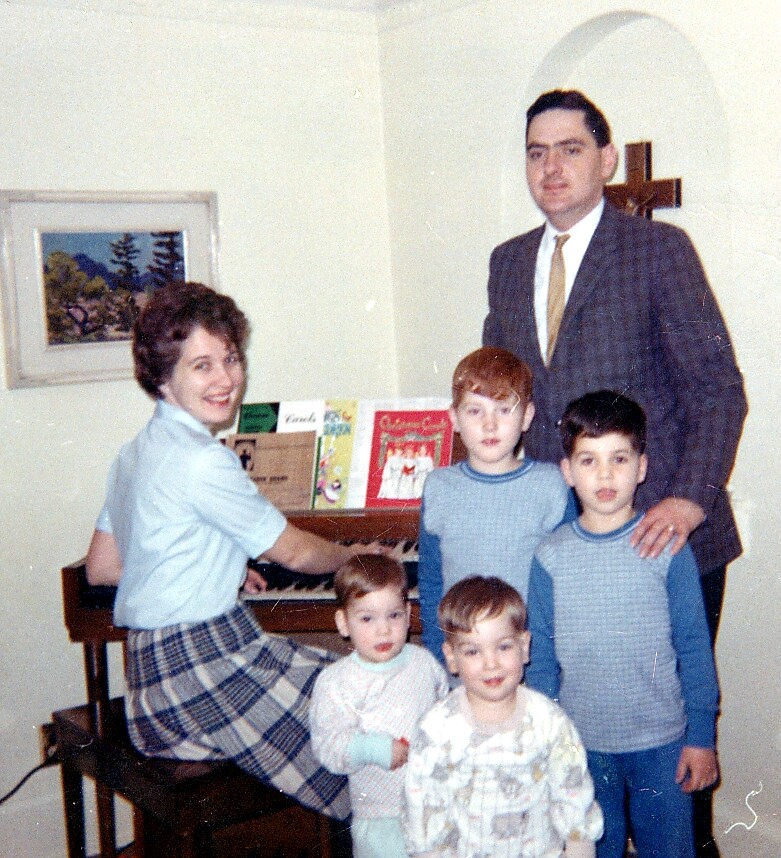 Family portrait in a living room: mother at the piano, father standing behind several children, cross on the wall in the background.