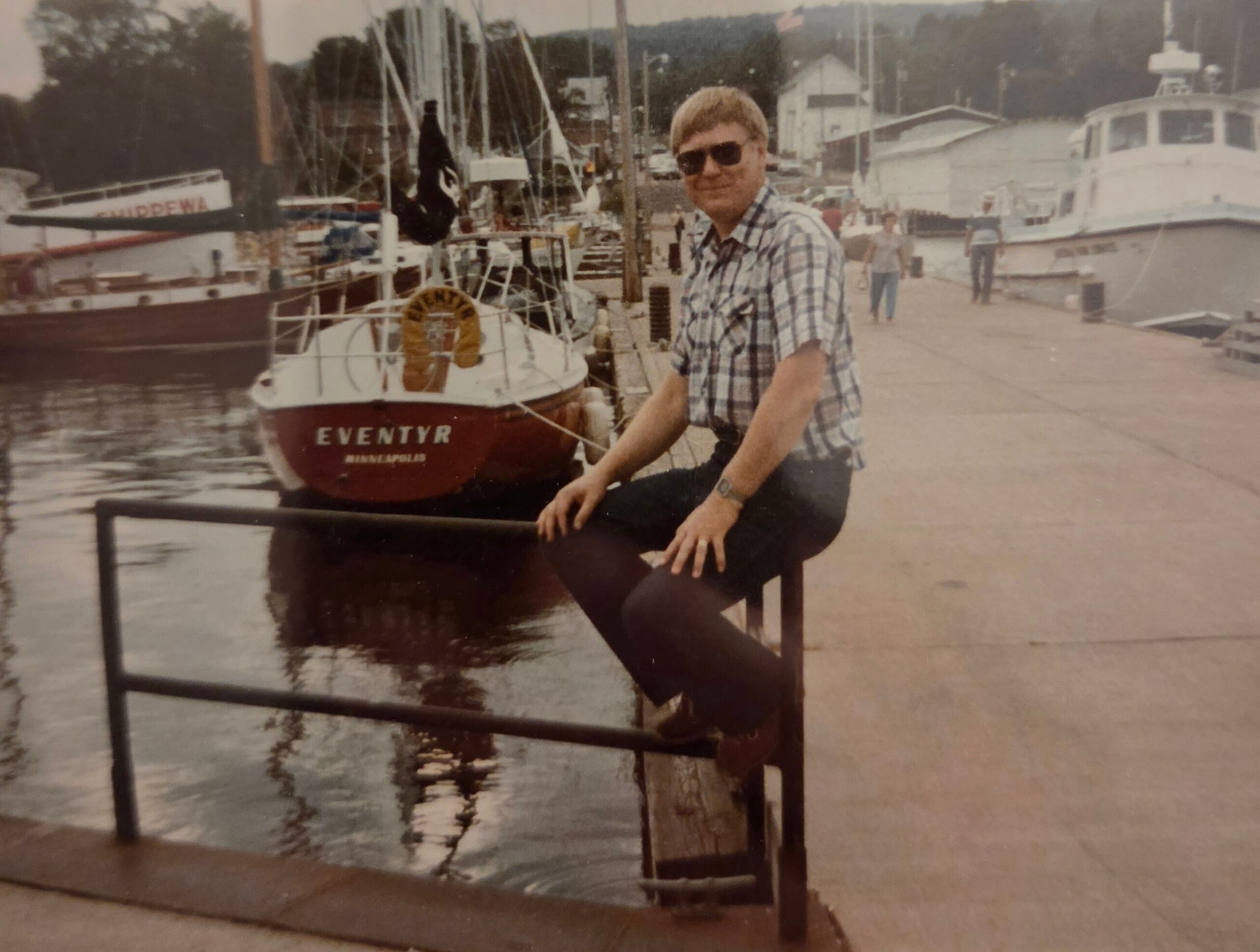 Man wearing sunglasses and a plaid shirt sits on a railing at a marina with boats docked in the background (vintage photo).