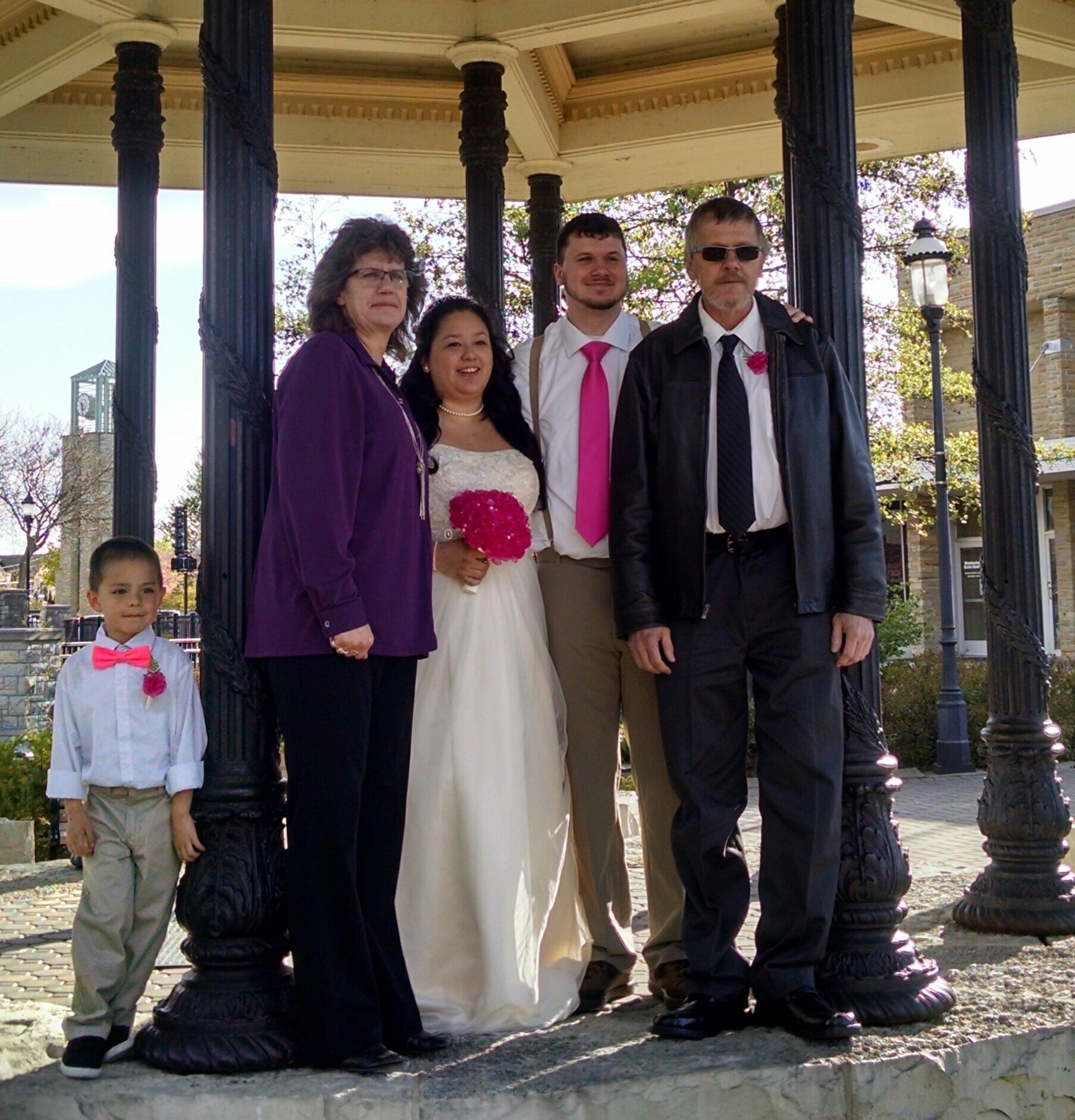 Bride in a white wedding gown holding a pink bouquet stands with four family members under a decorative gazebo on a sunny day.