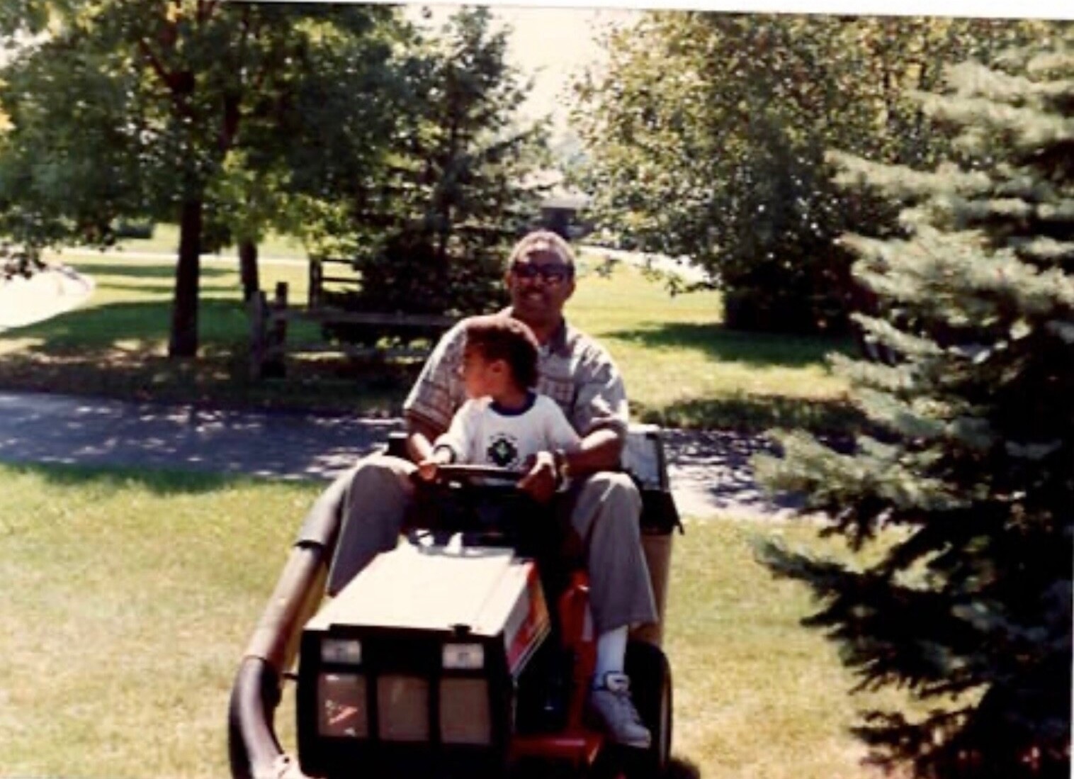 Man and child ride a red riding lawn mower in a sunny park, with trees and grass in the background, the adult supervising