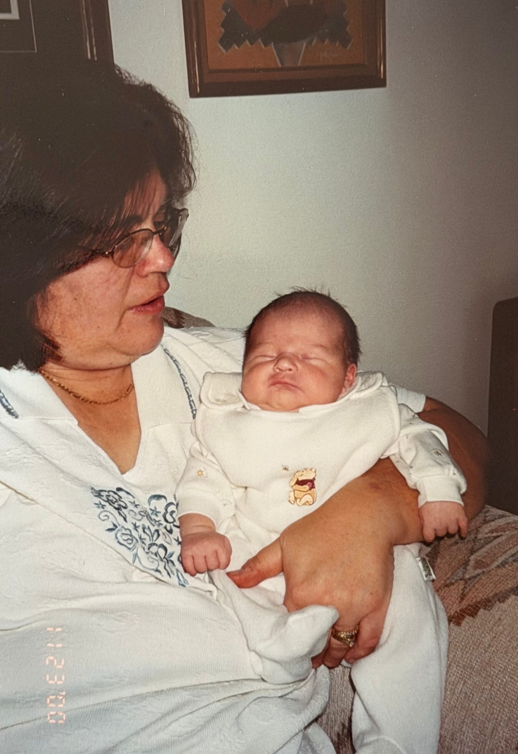 Older woman with glasses holding a newborn baby wrapped in white on a sofa at home.