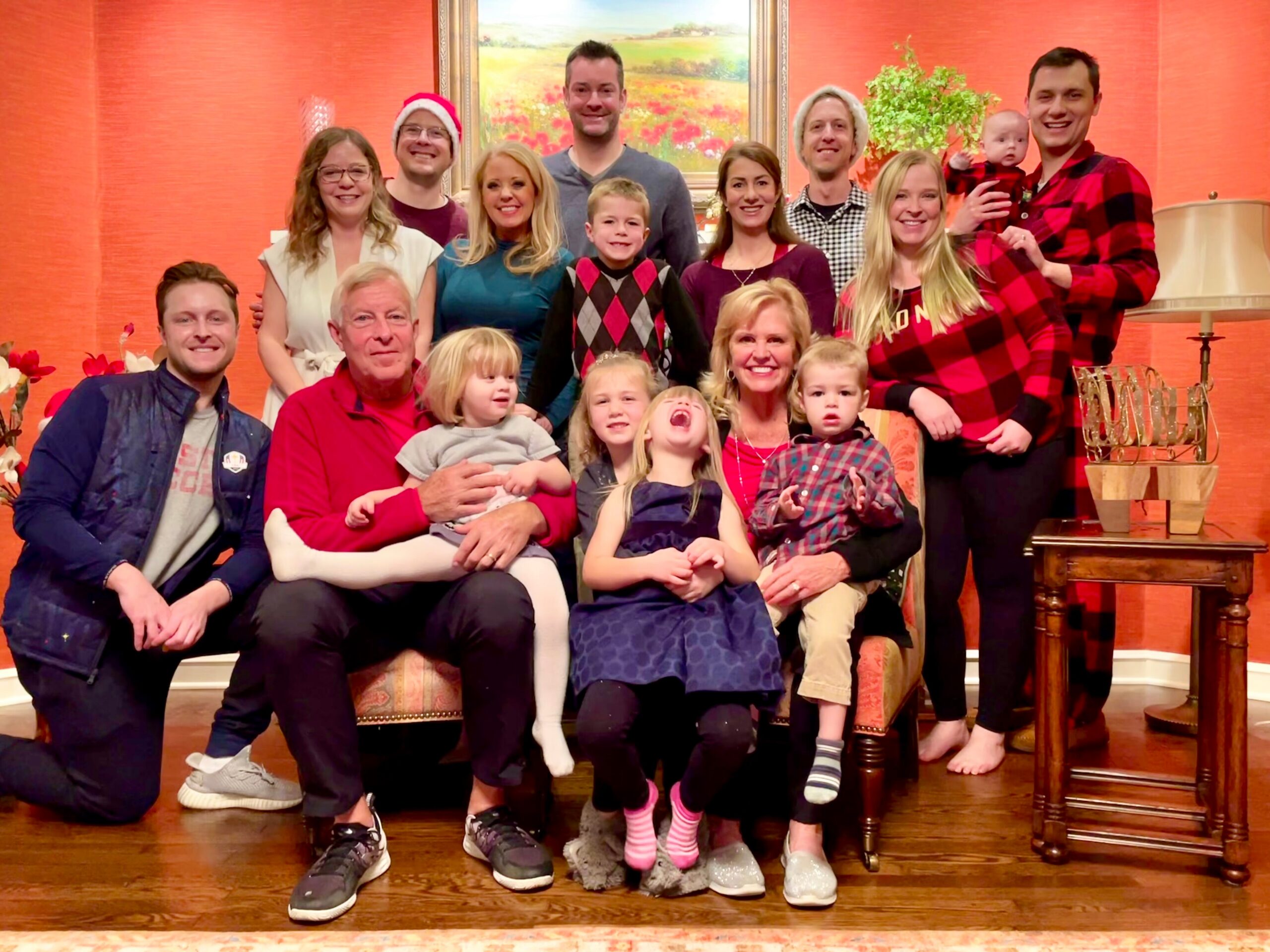 Large family group photo indoors: adults and children smiling, some in festive clothing, posed in a living room with an orange wall and a painting.