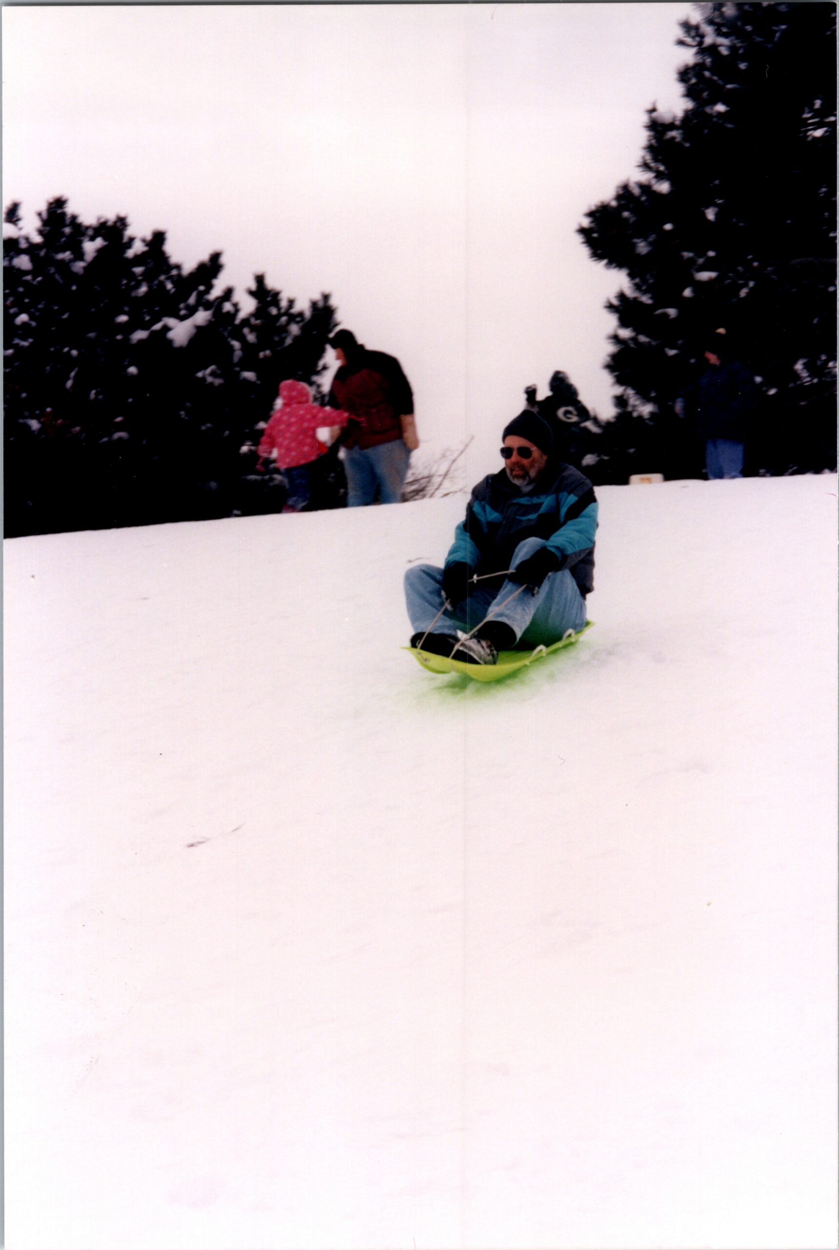 Man wearing a teal and black jacket sledding down a snowy hill, with a child and adults in the background near trees.
