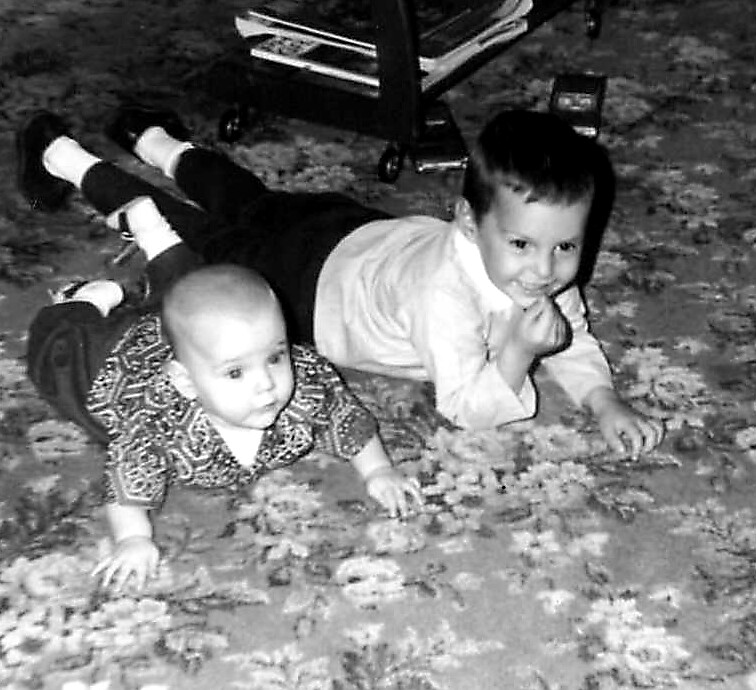 Two babies crawl on a flower-patterned carpet; one smiles at the camera while the other looks ahead.” John-style?