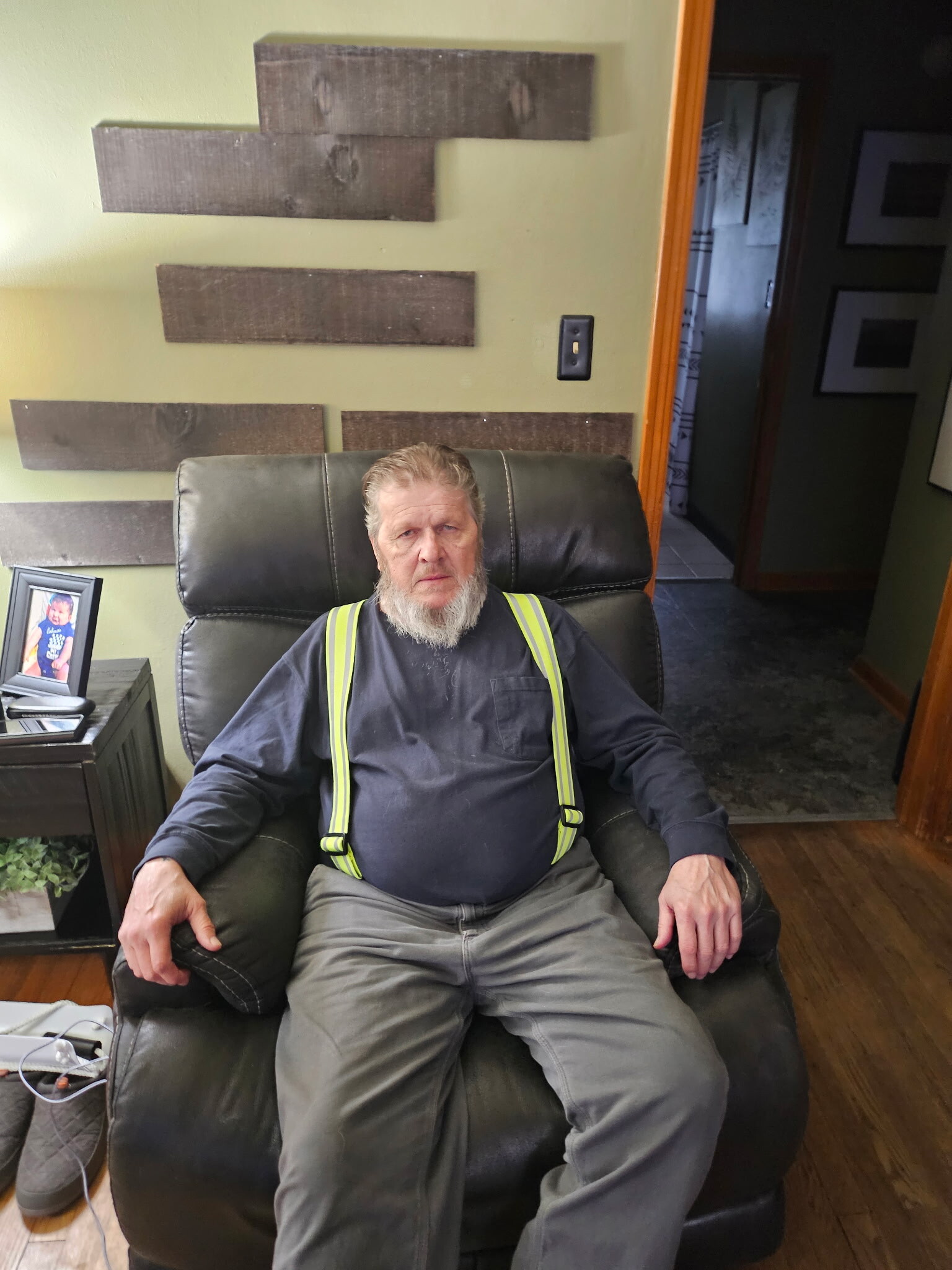 Older man with a gray beard sitting in a black leather chair, wearing a dark shirt and neon-yellow suspenders in a living room with a green wall and wooden wall decor behind him.