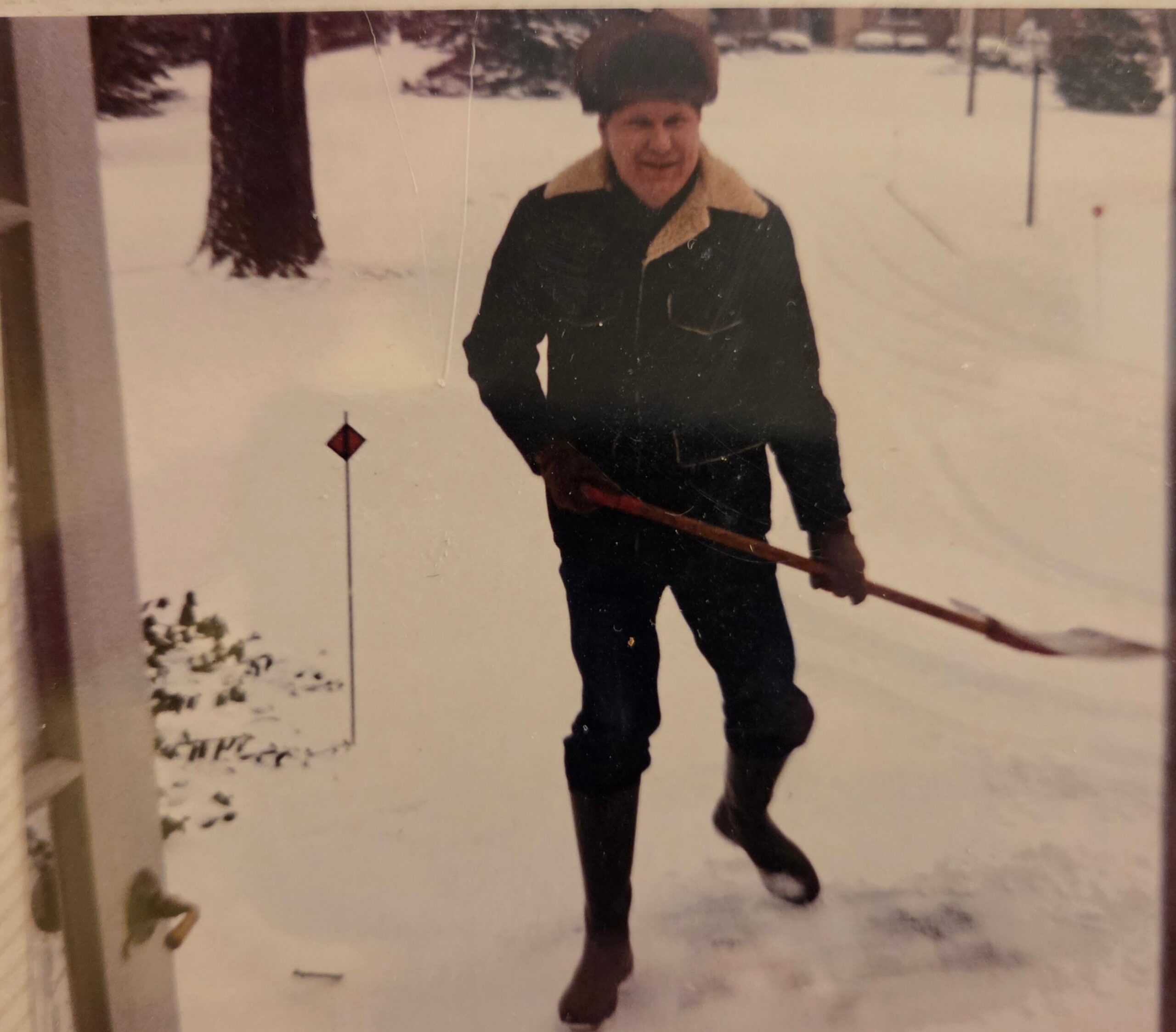 Man in a dark winter coat with a tan shearling collar and fur hat stands in deep snow, holding a long wooden ski pole or stick.